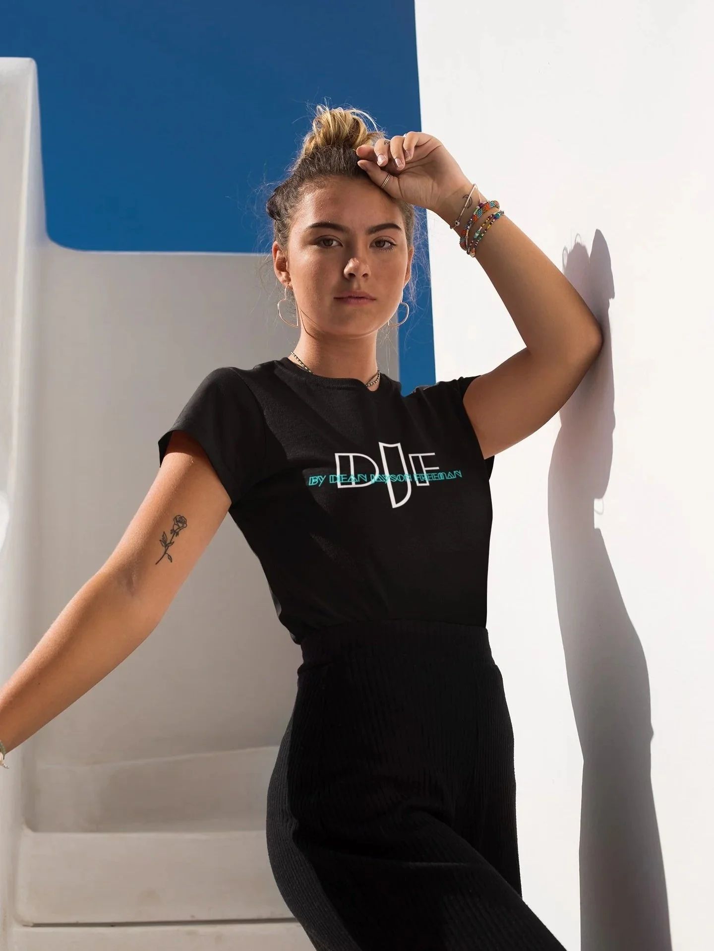 Junge Frau steht an einer weißen Wand, hebt die Hand an den Kopf und schaut ernst in die Kamera, trägt ein schwarzes T-Shirt mit Neon-Schriftzug und schwarze Ripphosen.