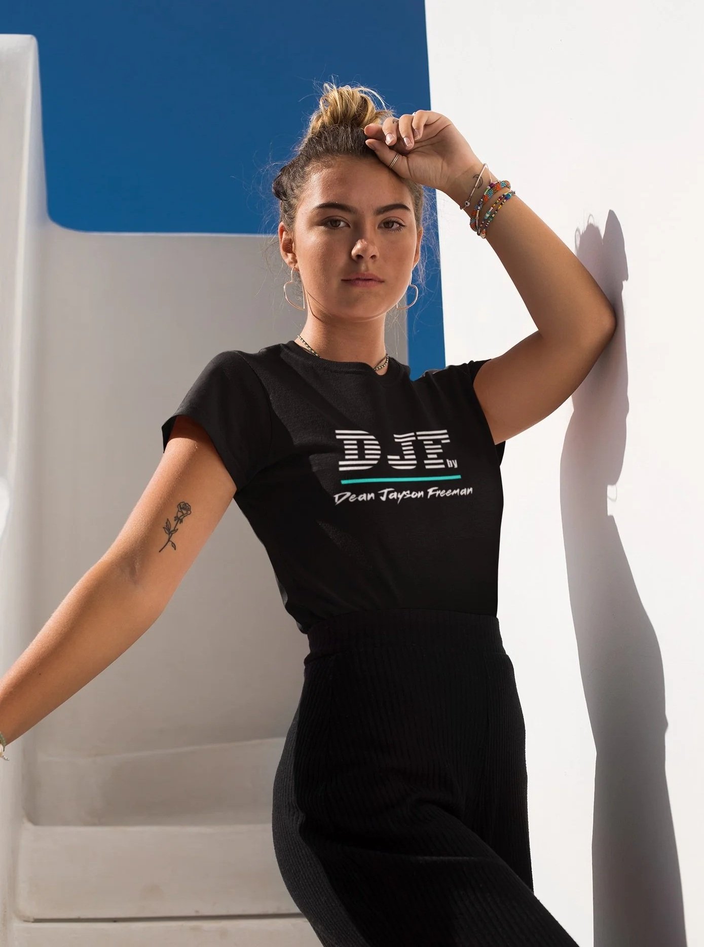 Junge Frau mit Tattoo auf dem linken Arm, trägt eine schwarze T-Shirt mit 'DJF' und 'Dean Jayson Freeman' Schriftzug und schwarze Hose, steht vor einer weißen Wand mit blauer Ecke, hebt eine Hand zur Stirn.