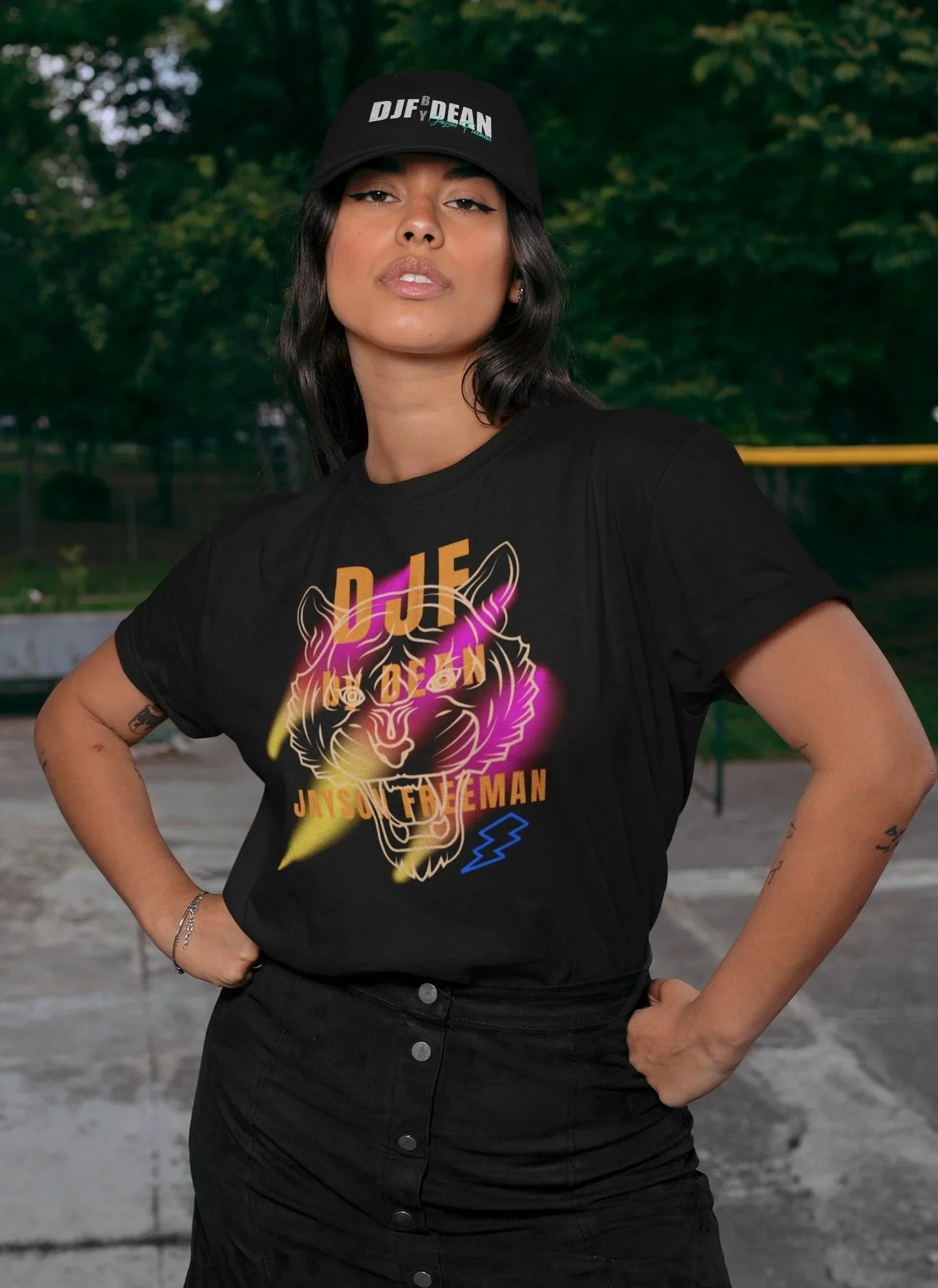 Junge Frau trägt schwarze T-Shirt mit leuchtendem Tiger-Design und Text, schwarze Cap, posed outdoor in der Nähe eines Basketballplatzes, grüner Hintergrund.