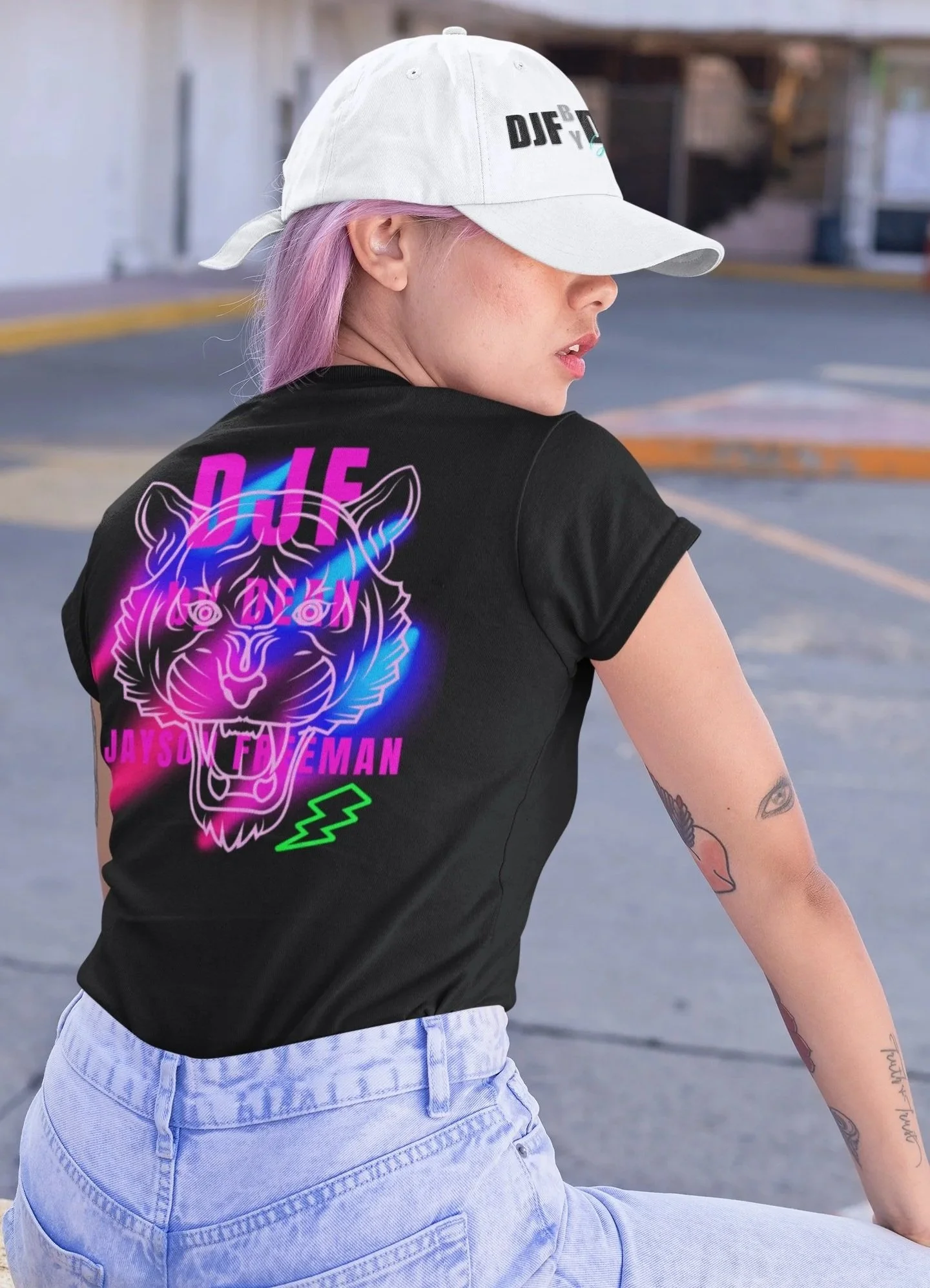 Junge Frau mit lila Haaren trägt schwarze T-Shirt mit Neon-Design und weißen Baseball-Cap auf einem Parkplatz.