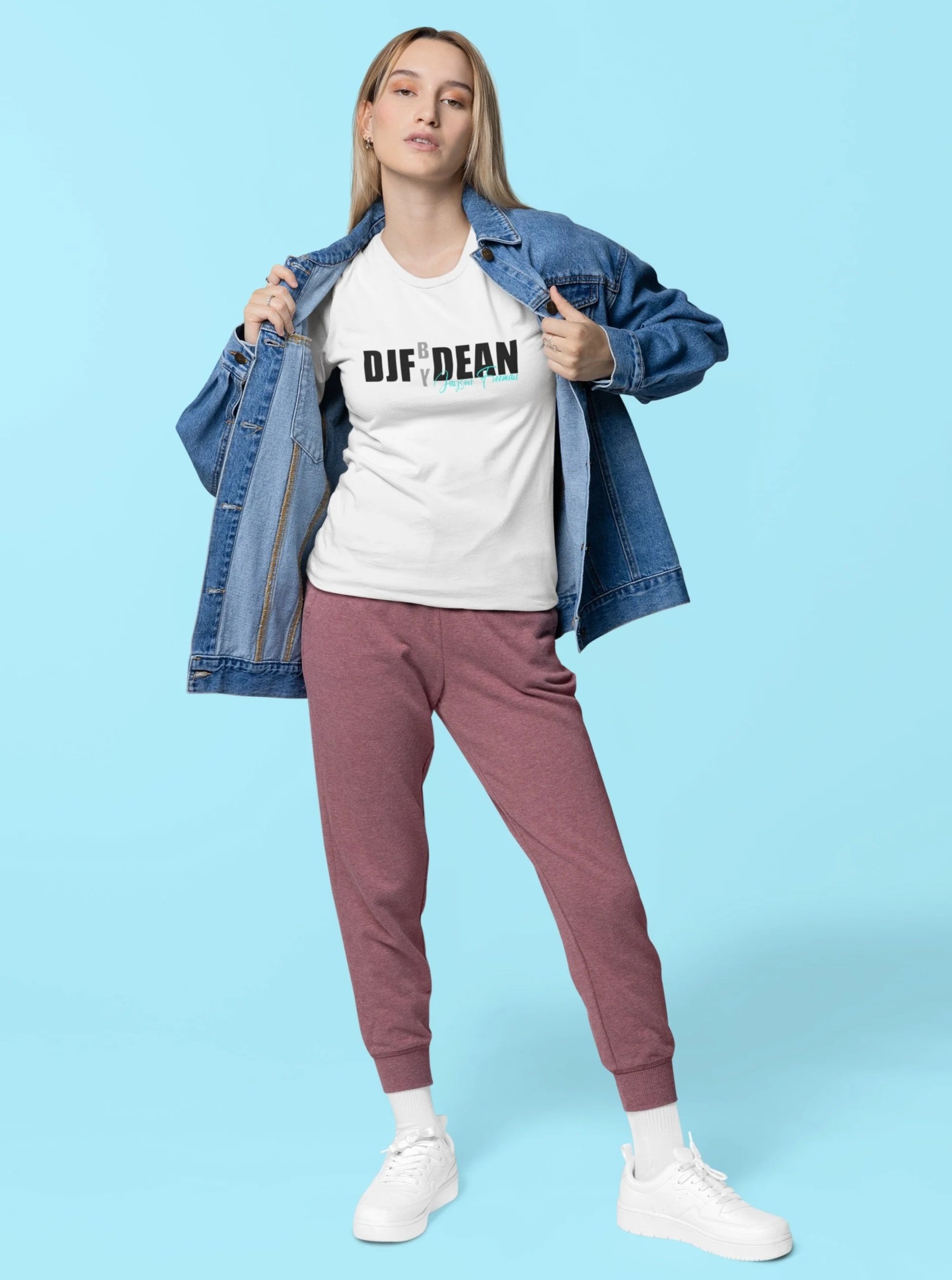 Eine junge Frau trägt einen weißen T-Shirt mit schwarzem Text, eine blaue Jeansjacke, pinkfarbene Jogginghose und weiße Sneakers, steht vor einem hellblauen Hintergrund.