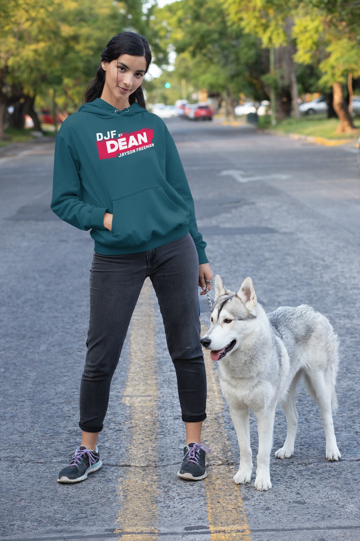 Junge Frau mit dunklen Haaren und grafischem Pullover steht auf einer Straße mit einem weißen Hund, umgeben von grünen Bäumen.