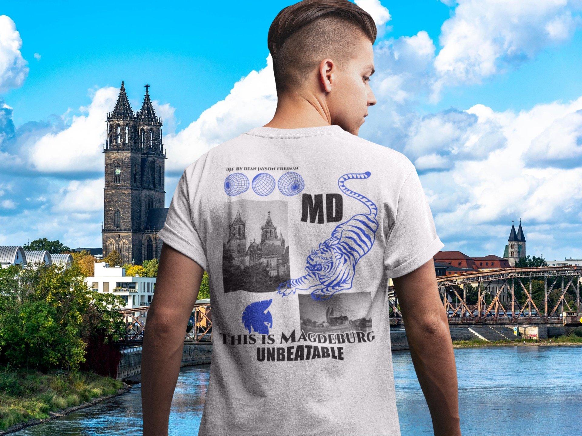 Ein junger Mann trägt ein weißes T-Shirt mit Aufdruck, im Hintergrund sieht man die Skyline von Magdeburg mit dem Dom und der Elbe bei schönem Wetter.