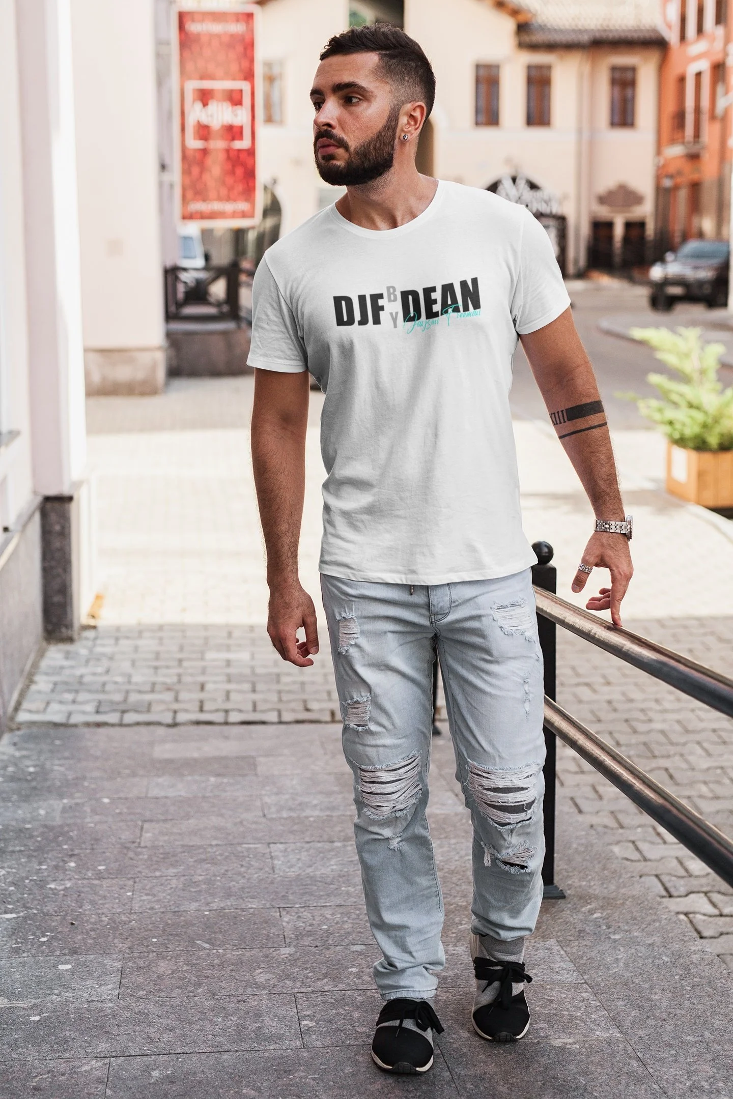 Junger Mann steht auf Gehweg in urbaner Umgebung, trägt weiße T-Shirt mit Aufdruck, zerrissene Jeans und Sneakers.