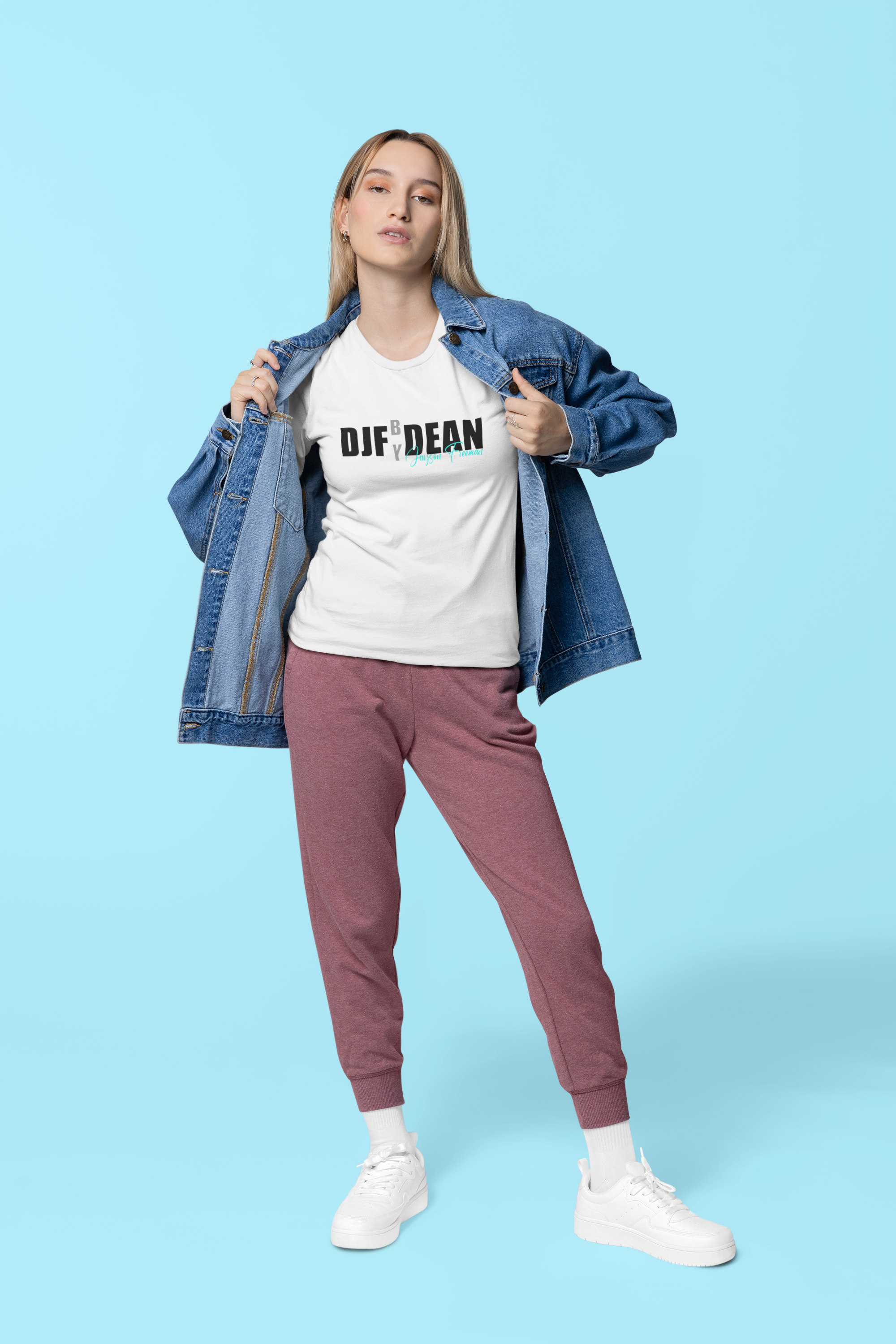 Junge Frau in Jeansjacke, T-Shirt mit Text, Jogginghose und weißen Sneakers steht auf hellem blauen Hintergrund.