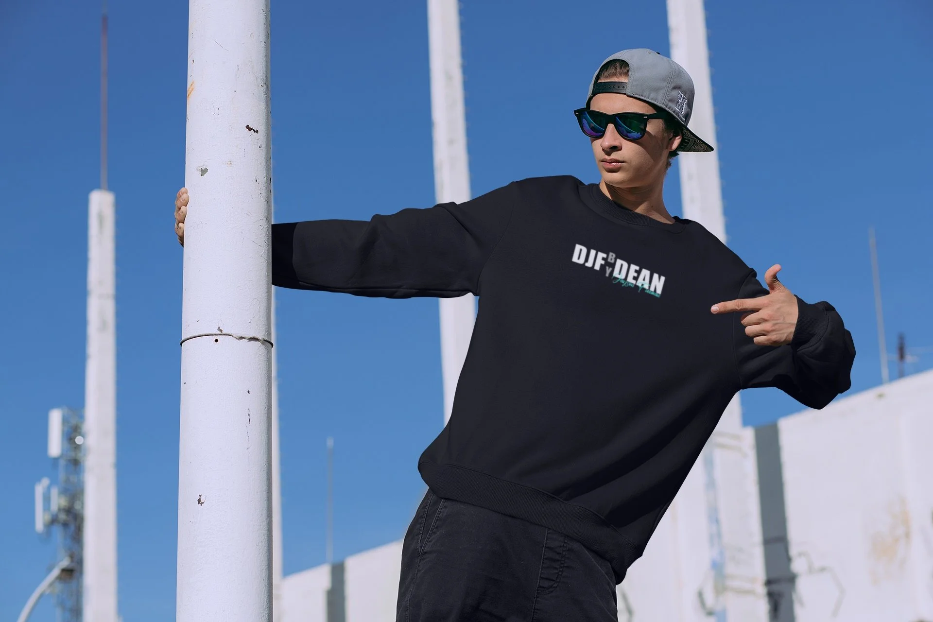 Junge Person trägt dunklen Pullover mit Aufdruck 'DJF BY DEAN', trägt Sonnenbrille und Kappe, steht im Freien bei blauer Himmel, zeigt auf den Pullover, hält sich an einem weißen Pfosten fest.