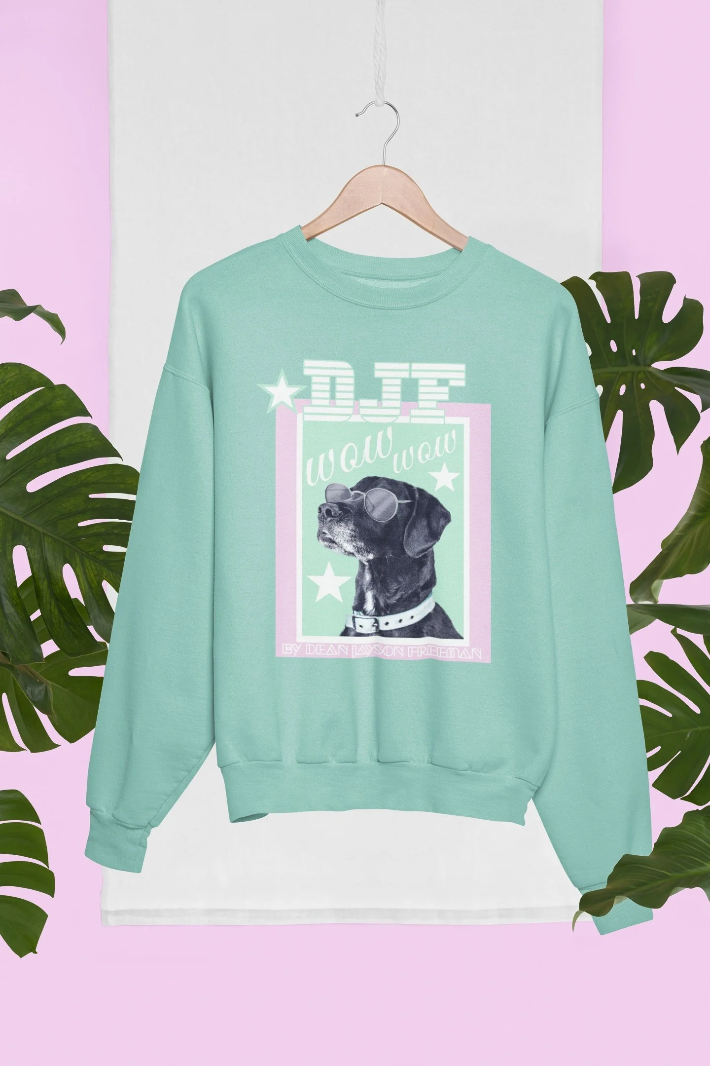 Ein mintgrüner Pullover mit einem Hund mit Sonnenbrille auf der Vorderseite, hängt an einer Holzkleiderstange vor einem weißen und lilafarbenen Hintergrund, umgeben von Monstera-Blättern.