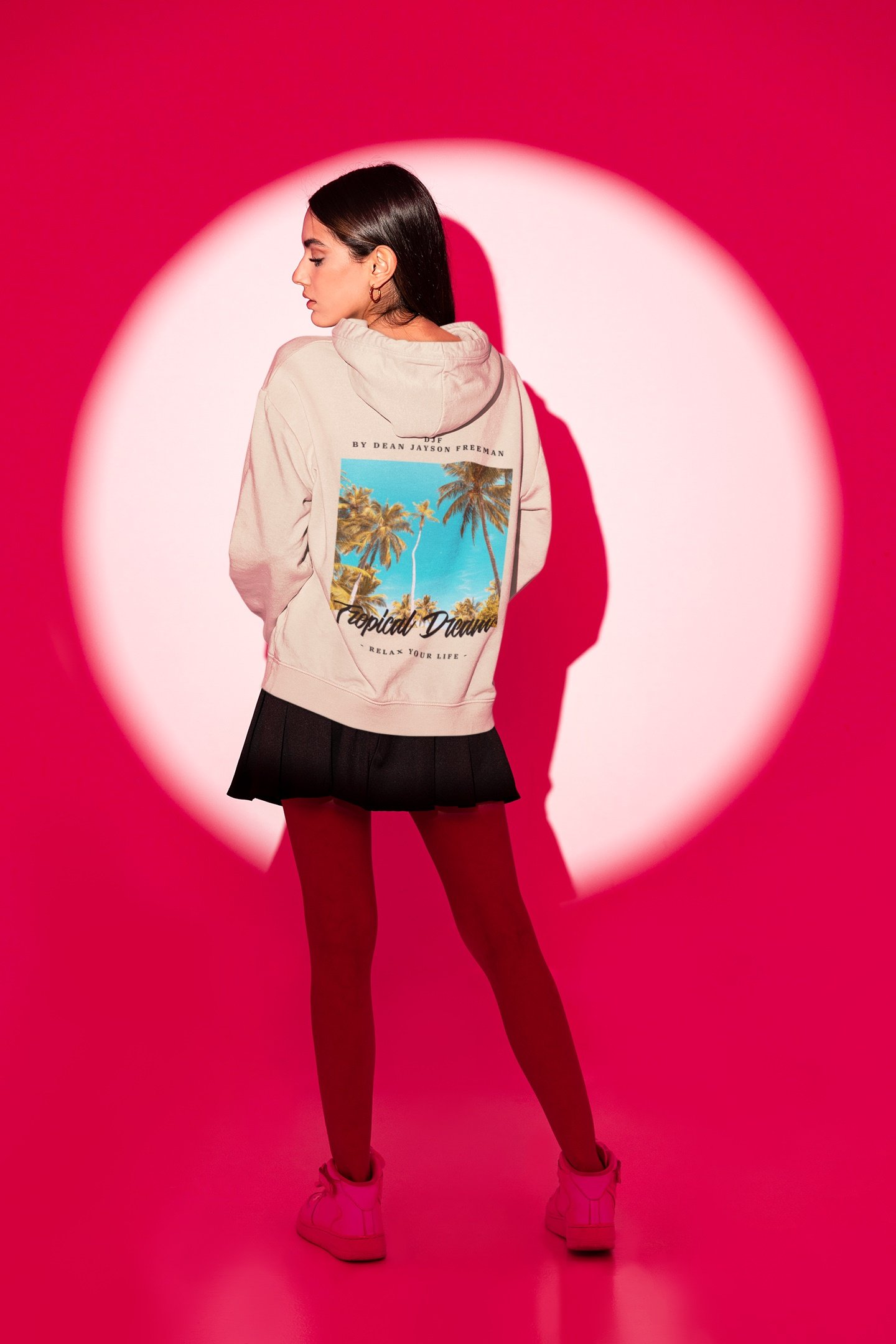 Junge Frau trägt Hoodie mit Strandmotiv, steht vor pinkem Hintergrund mit Spotlight-Licht, Beine verschränkt, wirkt nachdenklich.