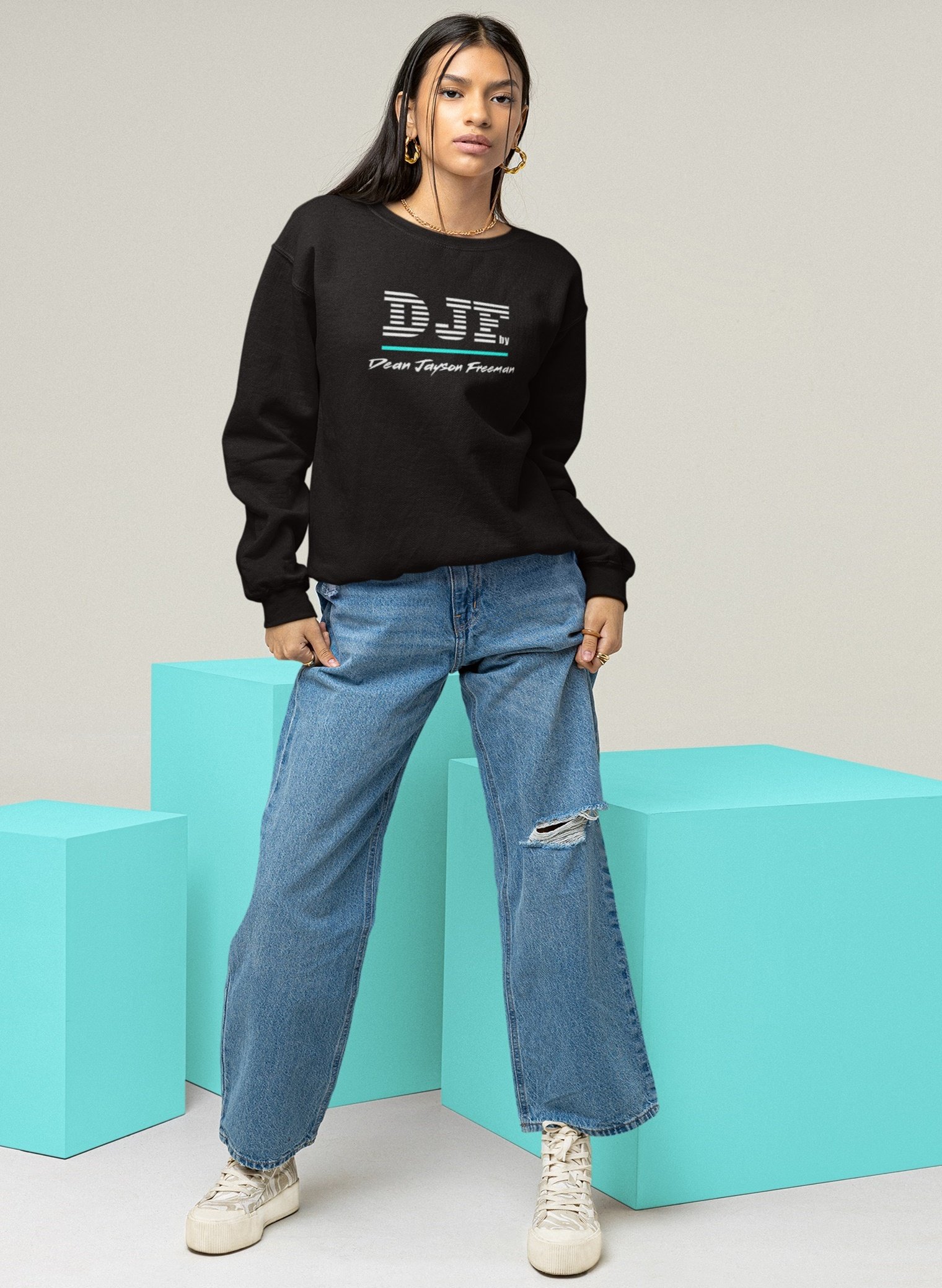 Junge Frau in schwarzem Sweatshirt und weiten, zerrissenen Jeans, steht vor blauen Quadern, trägt weiße Turnschuhe und große goldene Ohrringe.