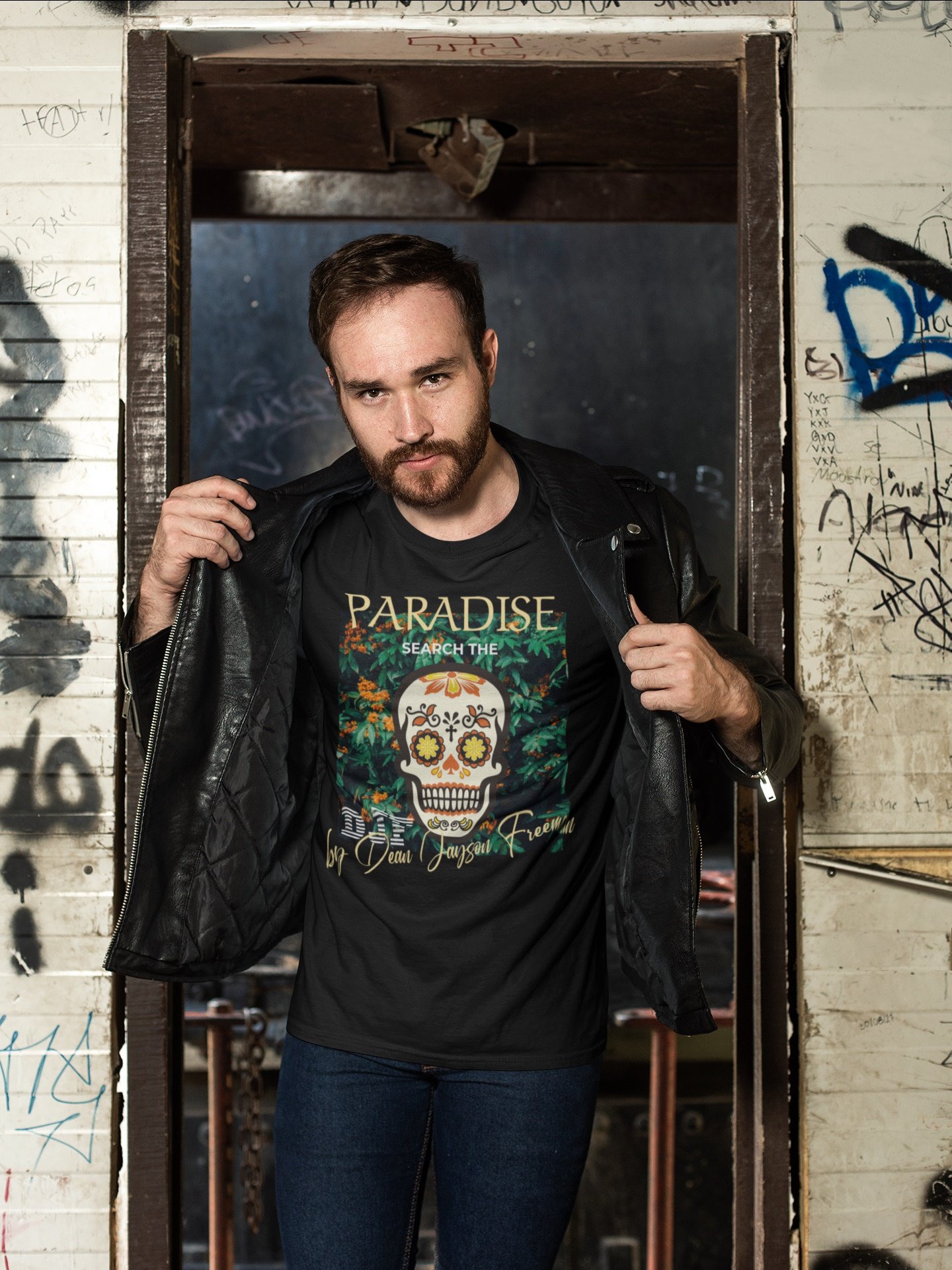 Ein Mann mit braunem Bart und kurzen braunen Haaren steht in einem graffitiüberwucherten, grauen Bereich und schaut direkt in die Kamera, während er eine schwarze Lederjacke trägt und eine schwarze T-Shirt mit einem bunten Totenkopf-Design anhat.