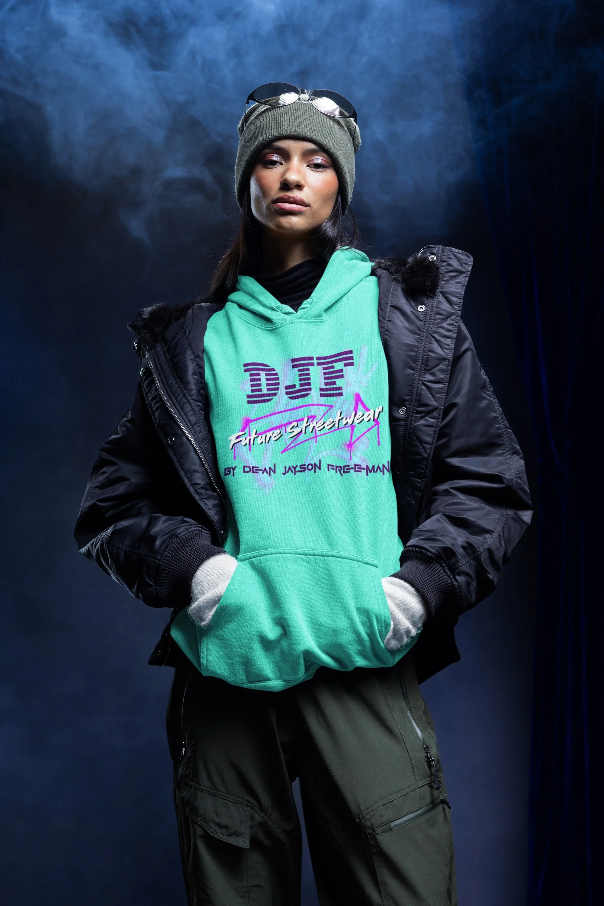 Eine junge Frau trägt eine graue Beanie, eine schwarze Jacke, eine grüne Hoodie mit buntem Aufdruck und eine schwarze Hose, steht vor einem dunklen Hintergrund mit Rauch.