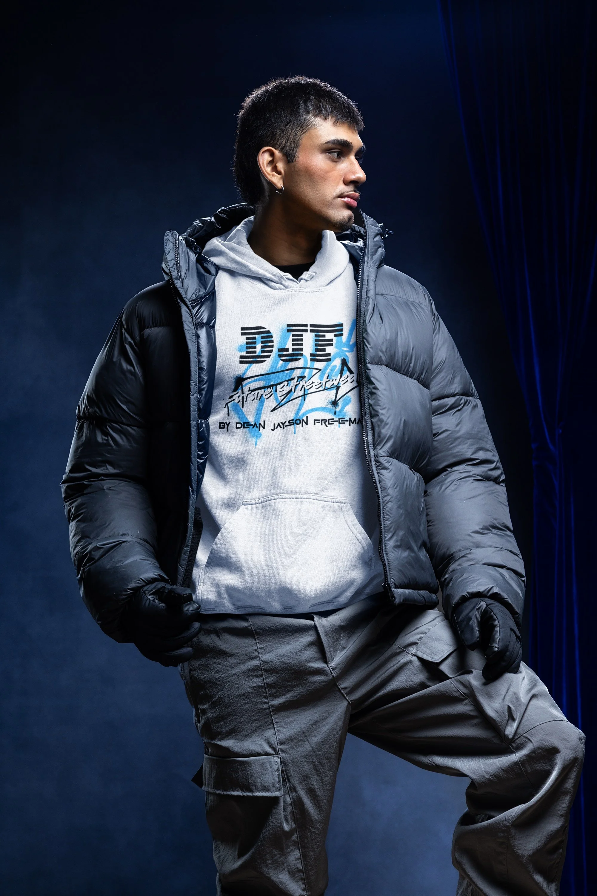 Junge Mann trägt eine graue Kapuzenjacke, eine weiße hoodie mit Schriftzug und graue Cargo-Hosen, vor dunklem, nebligem Hintergrund.