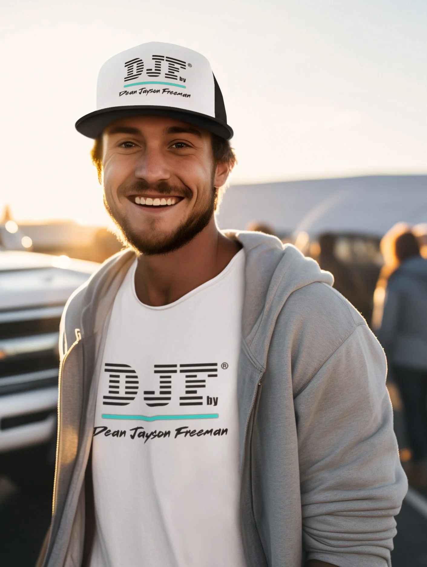 Lächelnder junger Mann trägt ein weißes T-Shirt und eine Mütze mit "DJE by Dean Tayson Freeman" auf einem Parkplatz bei Sonnenuntergang.