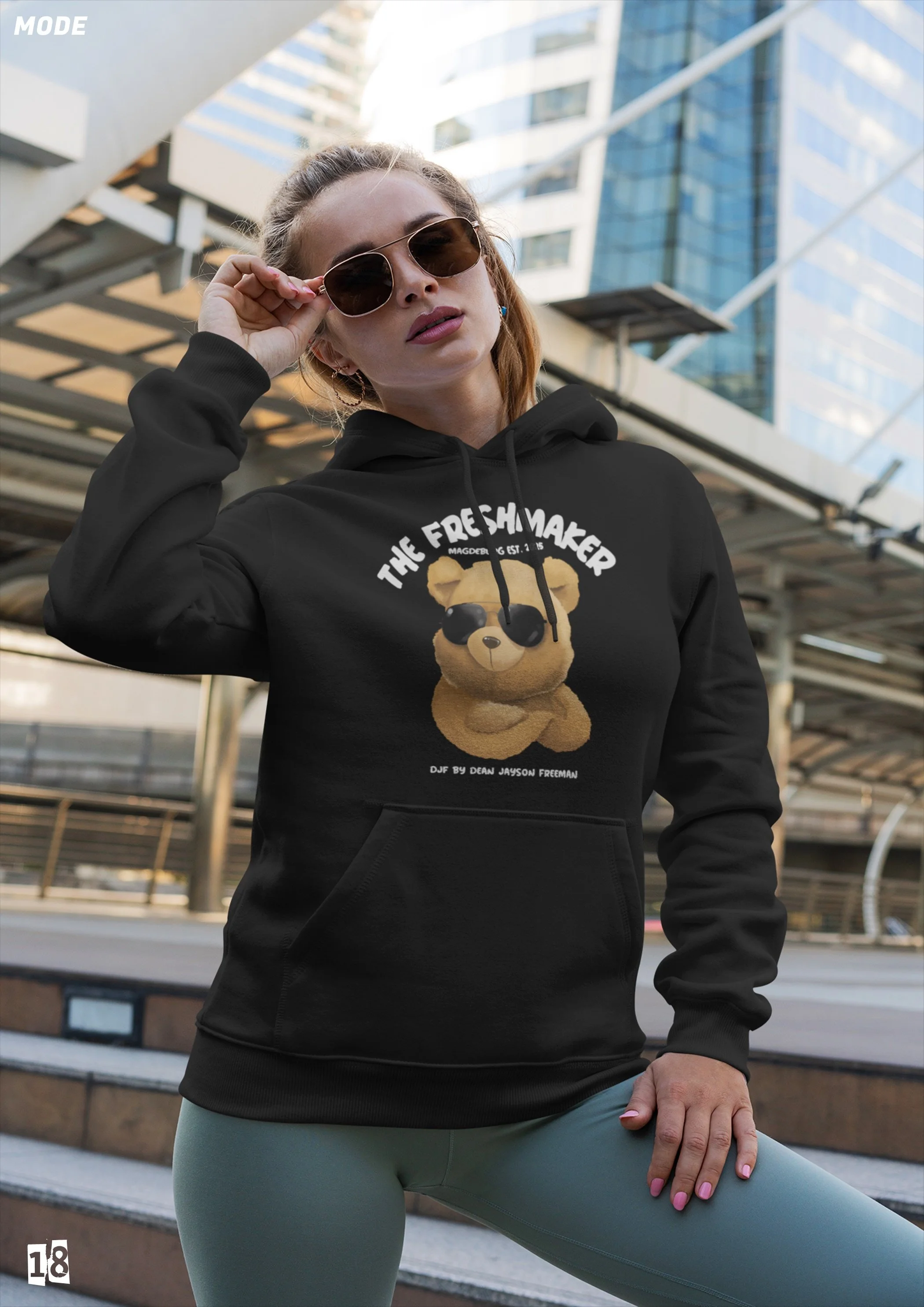 Frau in schwarzer Hoodie mit Teddy-Motiv und Sonnenbrille posiert vor modernen Gebäuden in der Stadt.