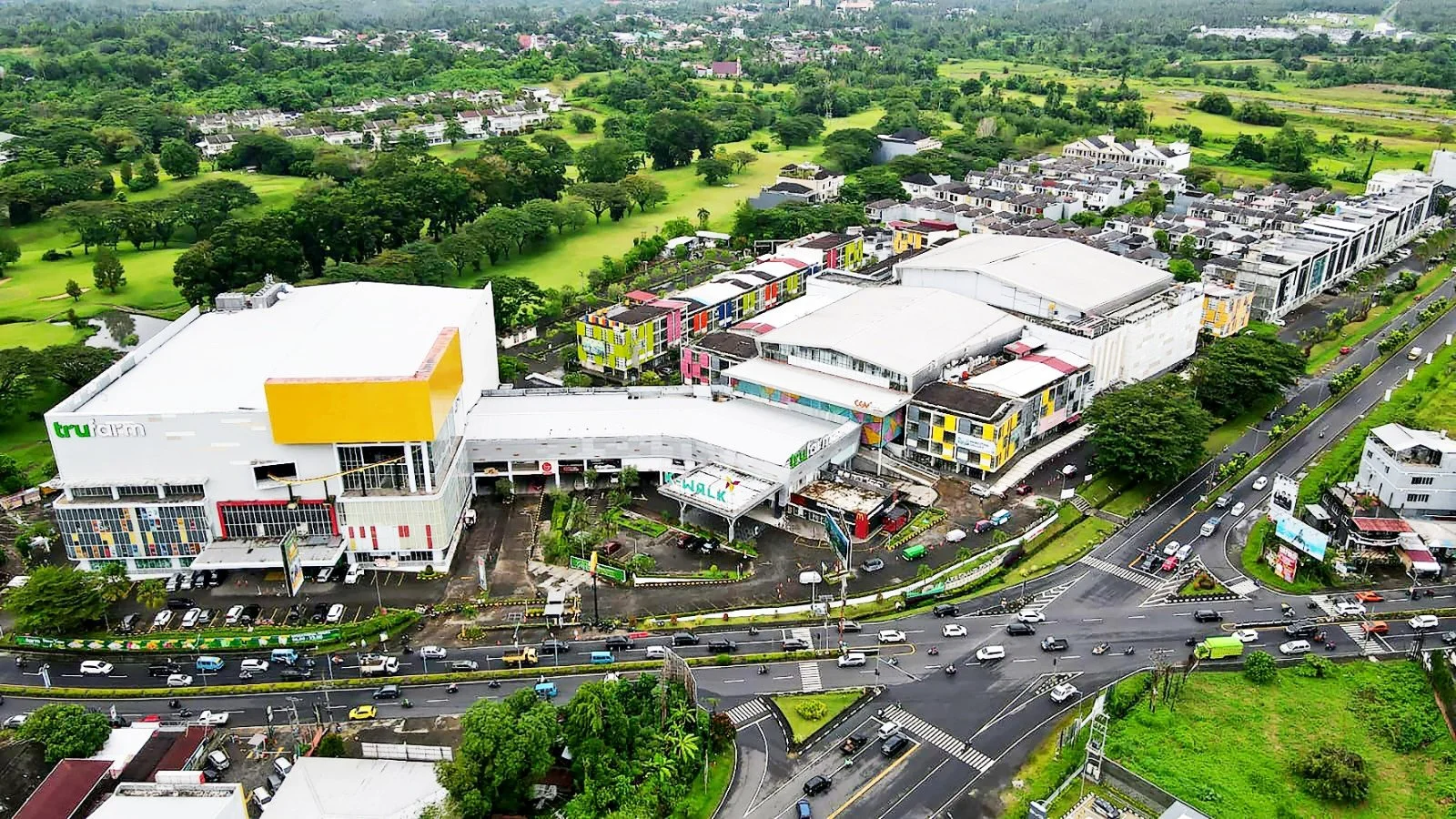 An aerial view of a large shopping mall with a parking lot, surrounded by trees and green landscape, with roads filled with cars in the foreground.
