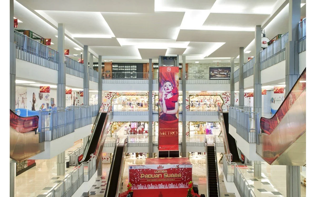 Interior of a multi-level shopping mall with escalators, railings, advertisements, and a large vertical banner of a woman in a red dress in the center.