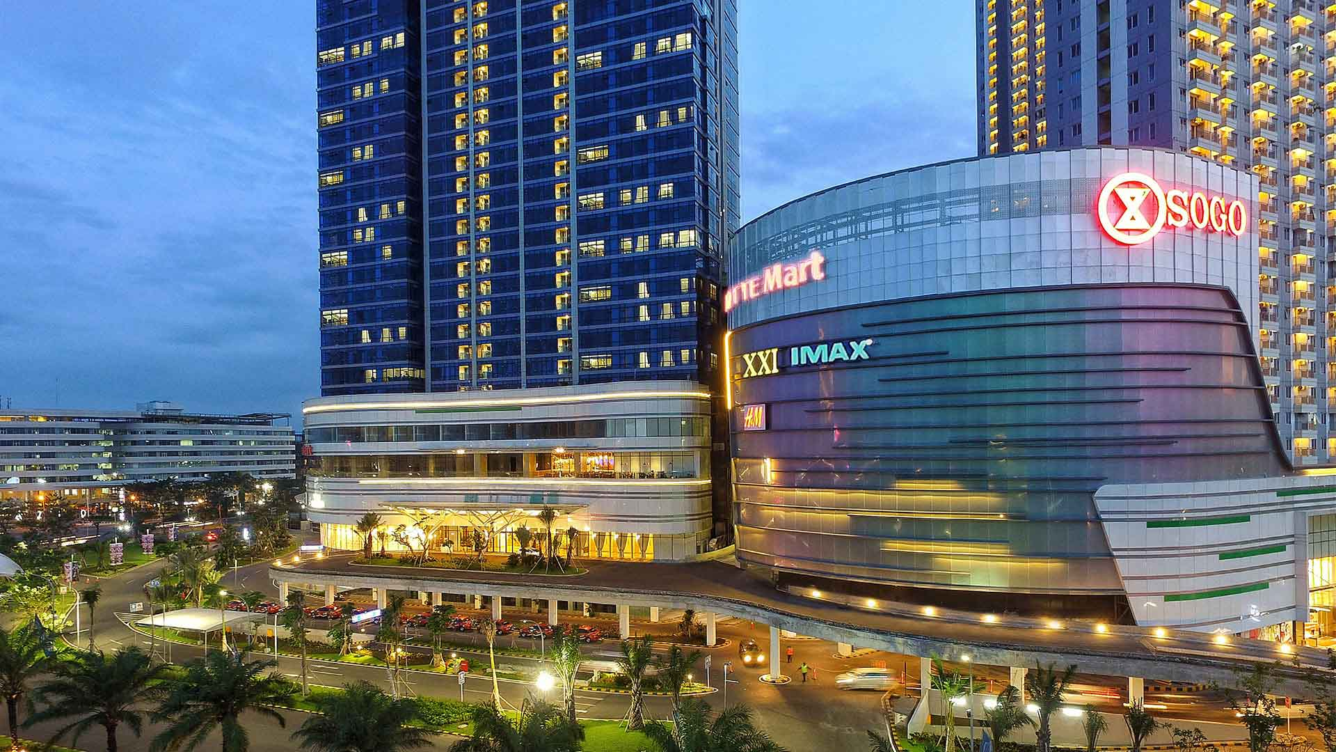 Nighttime view of a modern shopping mall with colorful neon signs, tall skyscrapers in the background, and a well-lit parking lot with cars and trees in the foreground.