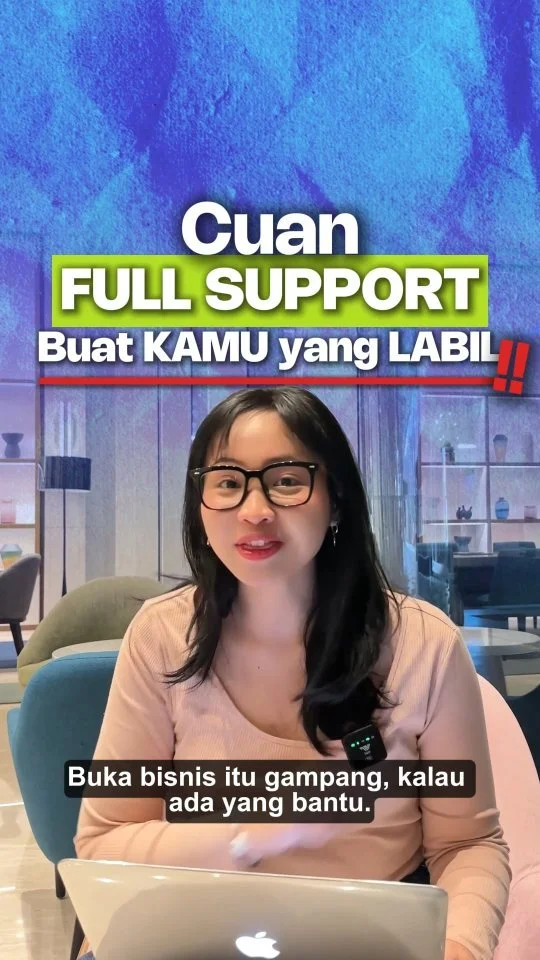 A woman with glasses and black hair sitting at a table with a laptop, speaking in front of a colorful sign that reads, 'Cuan Full Support Buat KAMU yang LABIL!!'