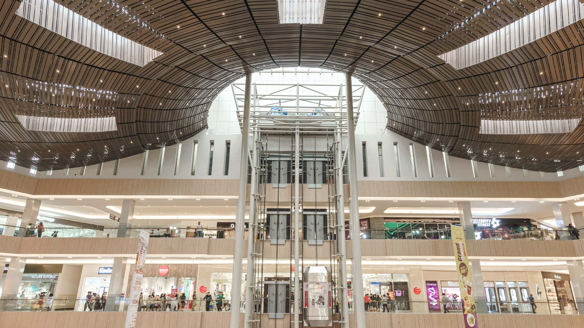 Interior of a modern shopping mall with multiple levels, a wooden ceiling with curved design, and stores with shoppers visible.
