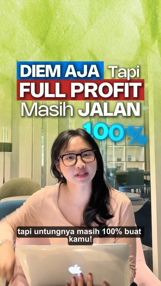 A woman with glasses and dark hair, sitting at a table with a silver MacBook, speaking in front of a colorful background with Indonesian text and a pen