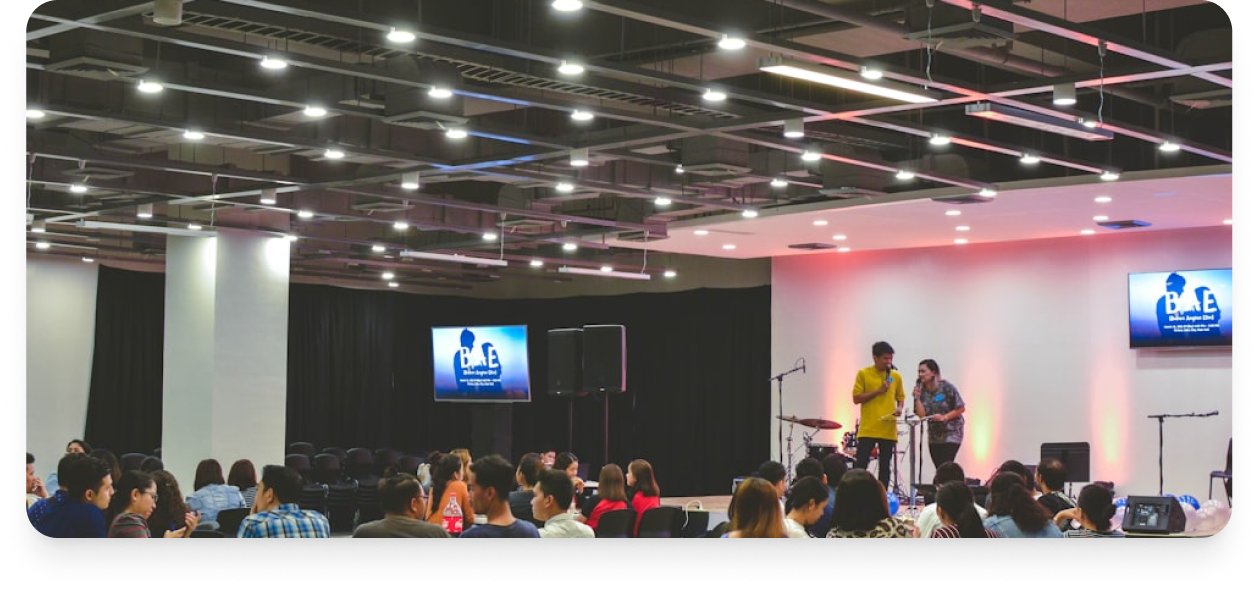 An indoor event with a stage, two people using microphones, and an audience seated, watching the performance with a screen displaying a logo.