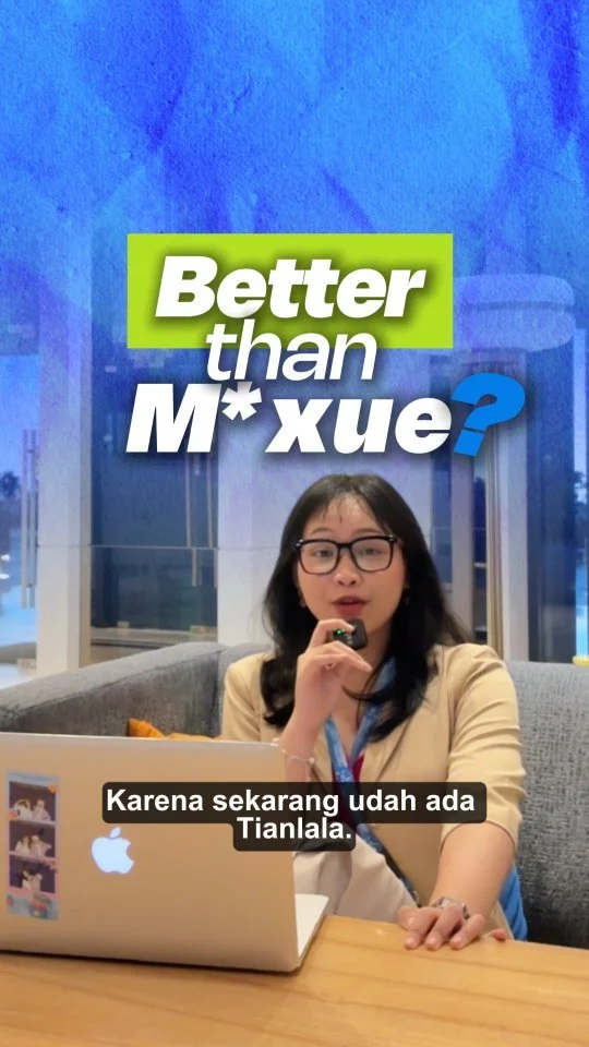 A woman with glasses and long dark hair sitting at a table with a laptop, in front of a colorful background that reads 'Better than M* xue?'. She is speaking into a microphone, and there are subtitles in Indonesian.