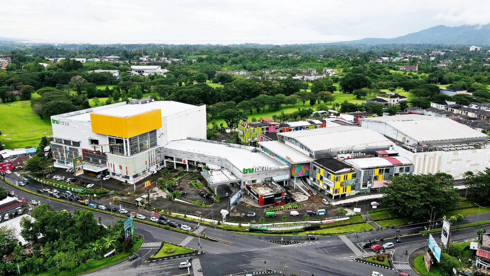 A large shopping mall with colorful building facades, surrounded by busy roads and green landscape in the background.
