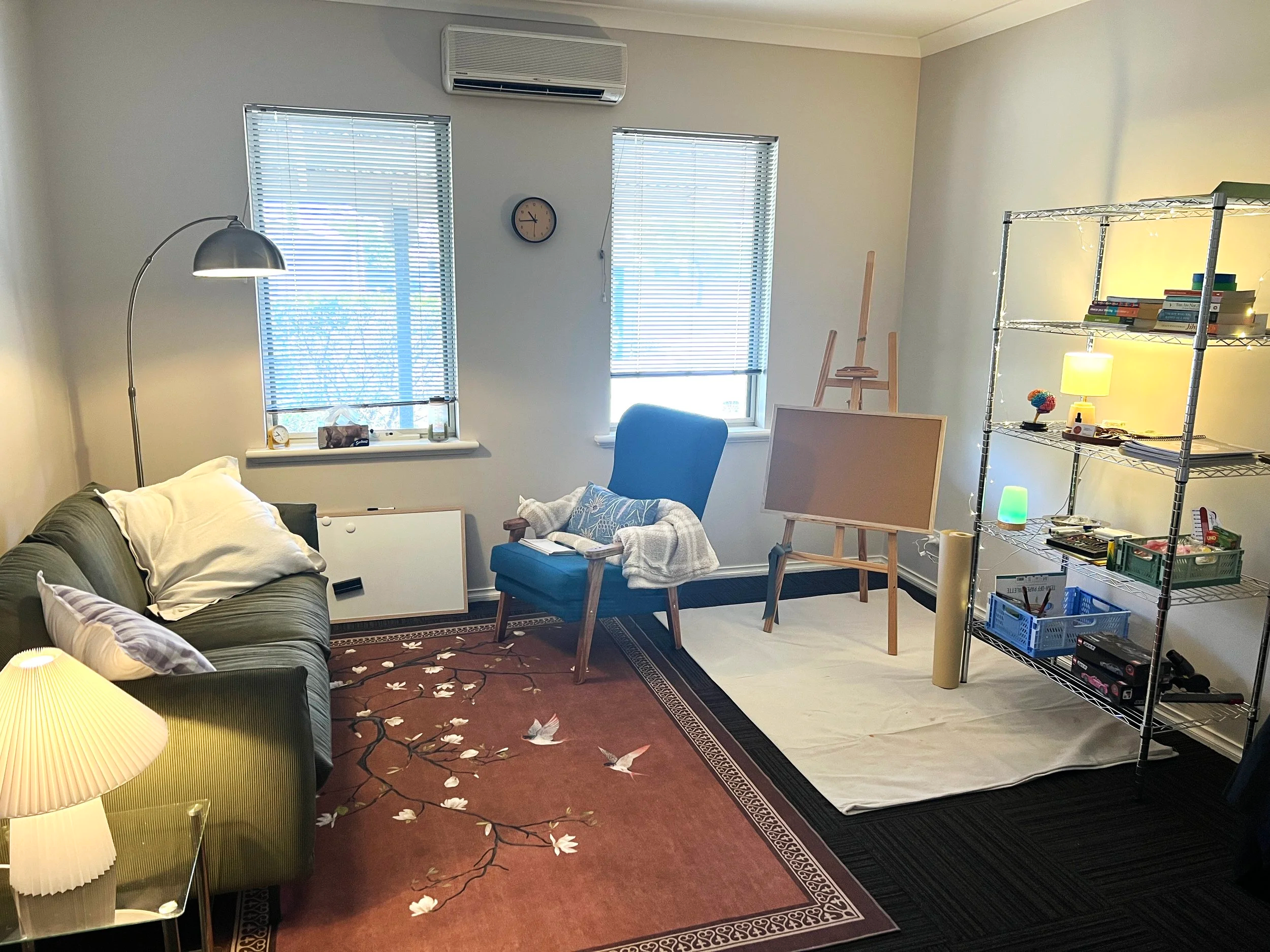 A cozy living room with two windows, a green sofa with pillows, a blue armchair with a book, an easel, a small white sheet on the floor, a metal shelving unit with books and lamps, a large rug with bird and flower designs, a floor lamp, a table lamp, and various decorative items.