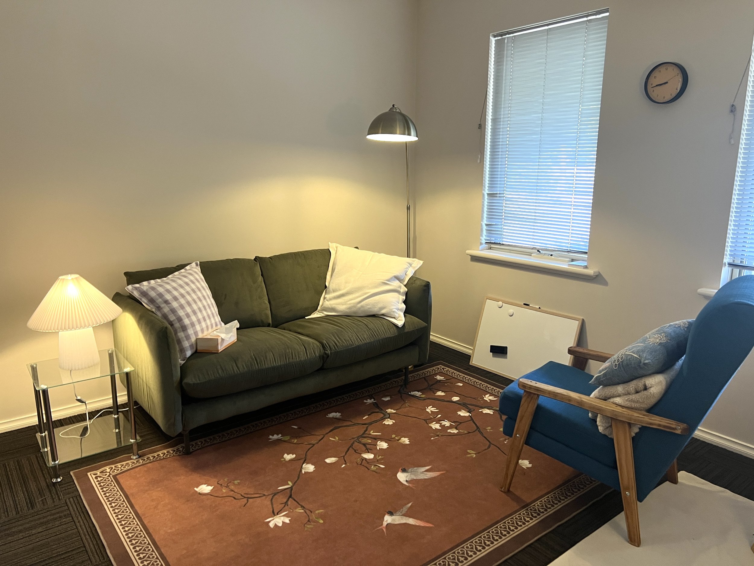 Living room with green sofa, blue armchair, side table with lamp, area rug with bird design, and windows with blinds.