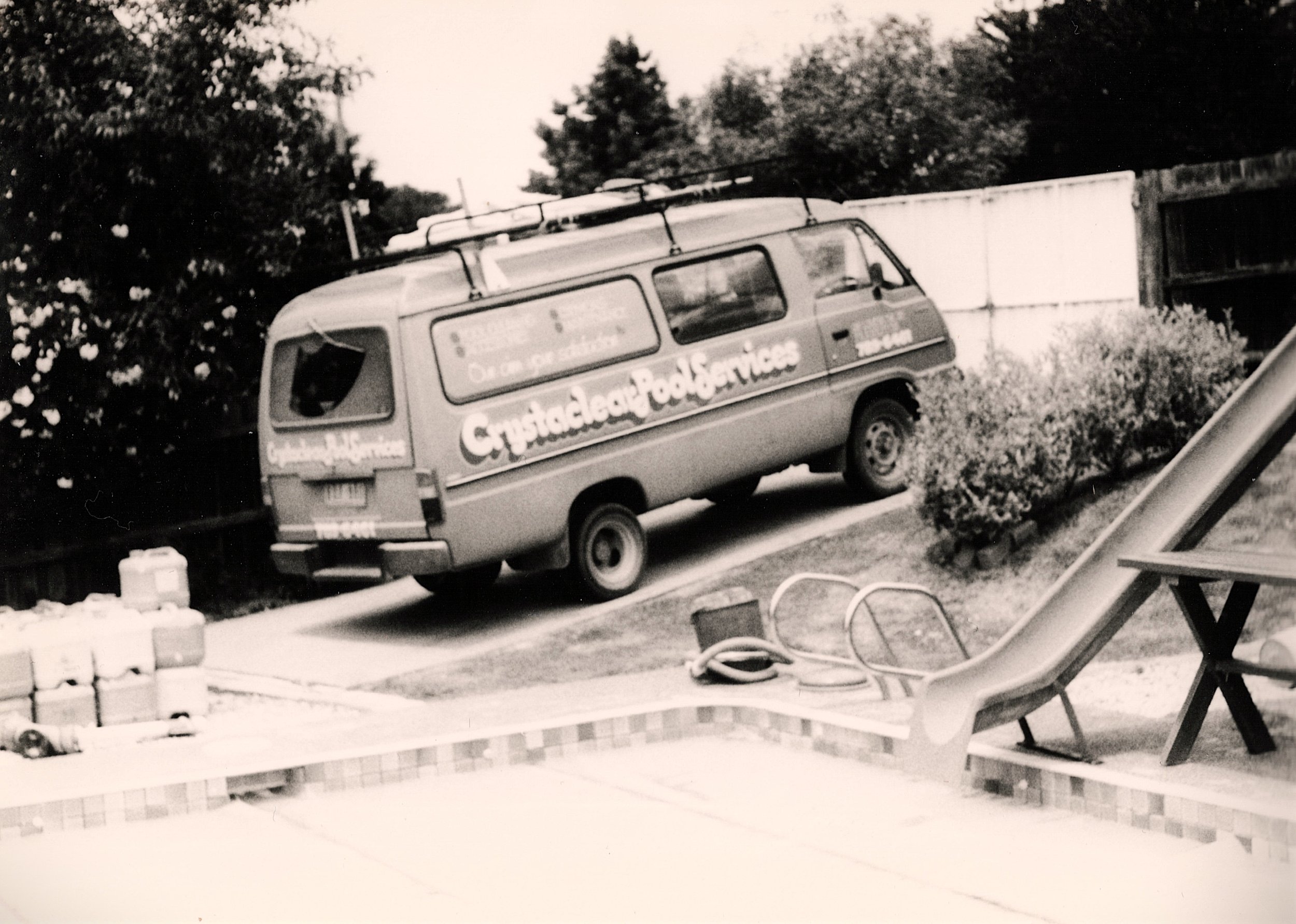 Black and white photo of a van parked in a yard with trees in the background. The van has signage advertising crystaclear pool services. There an old slide, covered swimming pool and chemical containers visible in the yard.