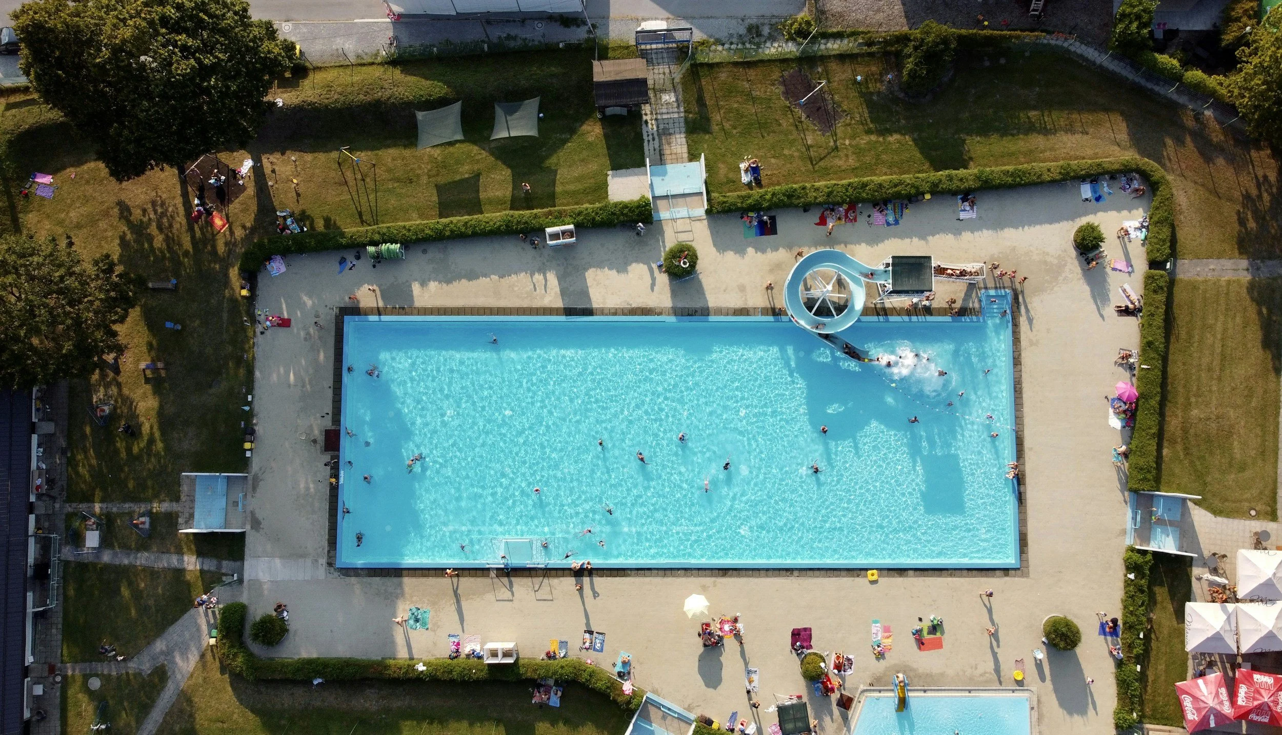 Aerial view of a swimming pool surrounded by a grassy area with trees, umbrellas, and people sunbathing and playing. There is a water slide in the pool with kids enjoying the water park area.