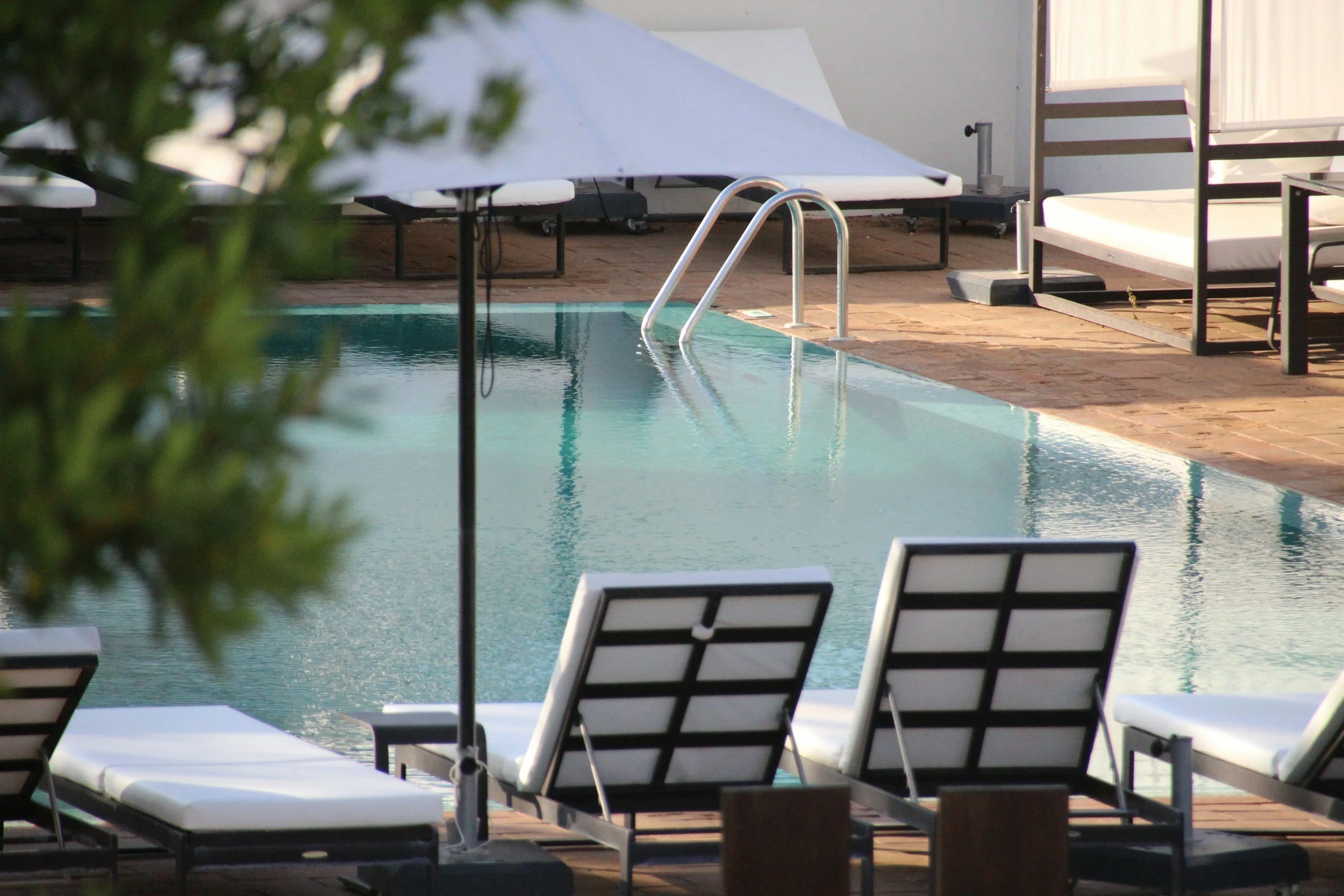 A swimming pool area with lounge chairs, umbrellas, and a canopy, surrounded by a brick patio and a white fence.