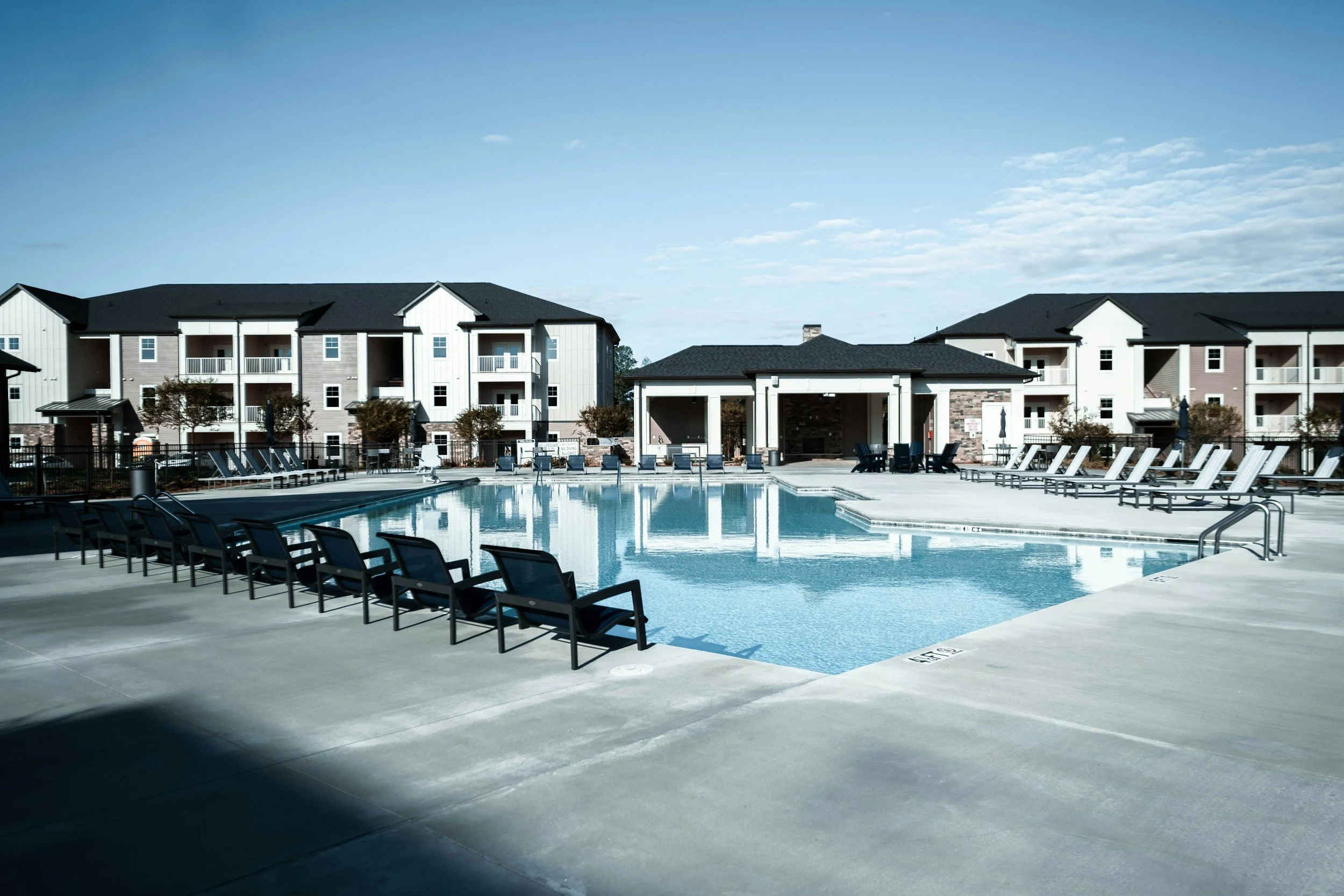 An outdoor swimming pool with lounge chairs along the poolside, surrounded by a fence, with apartment buildings in the background under a blue sky with some clouds.