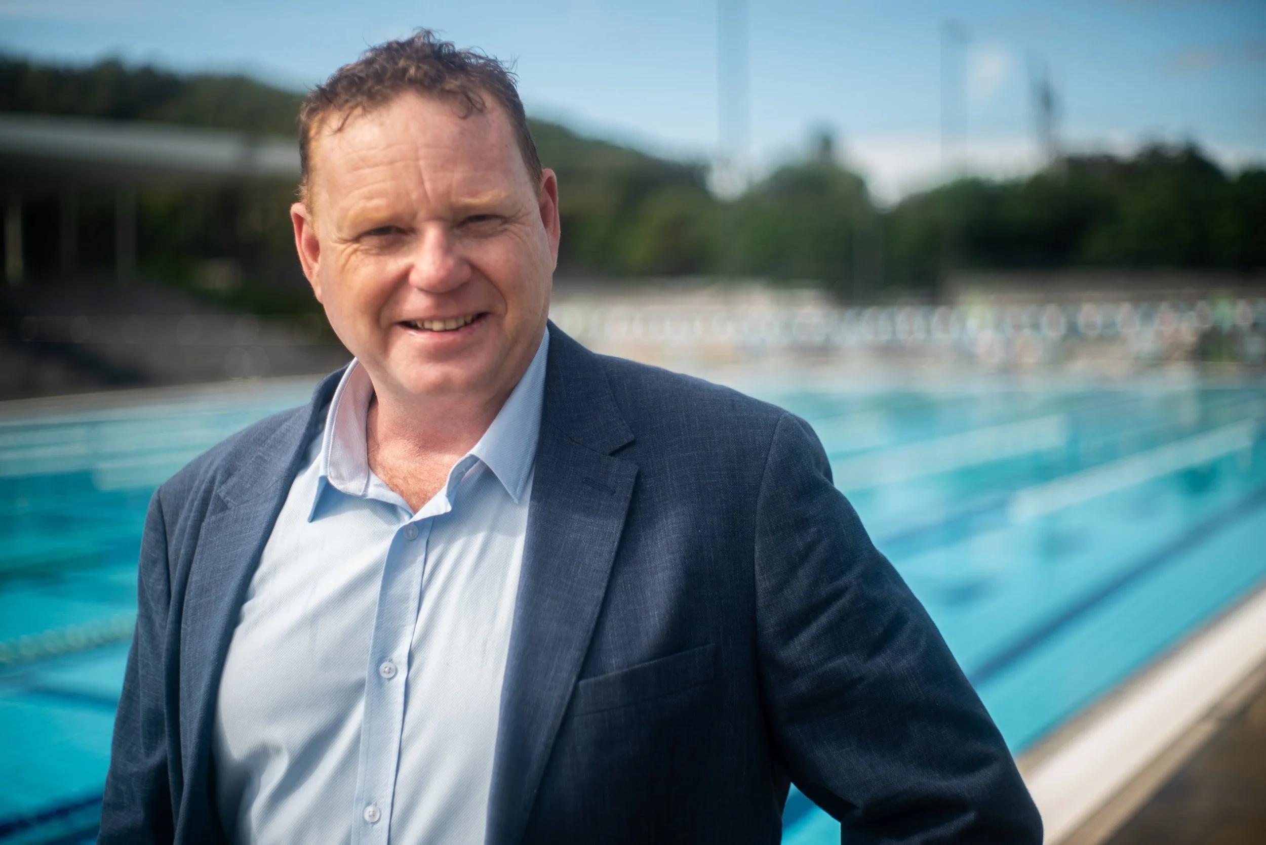 A man in a suit smiling outdoors near a swimming pool with a blurred background of trees and a fence. Nick Briscoe