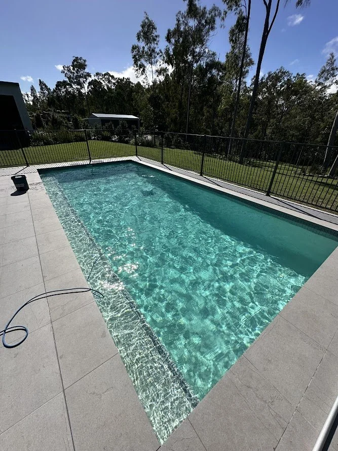 Empty swimming pool with clear water, surrounded by a concrete deck and black metal fence, with a green lawn and trees in the background under a partly cloudy sky.