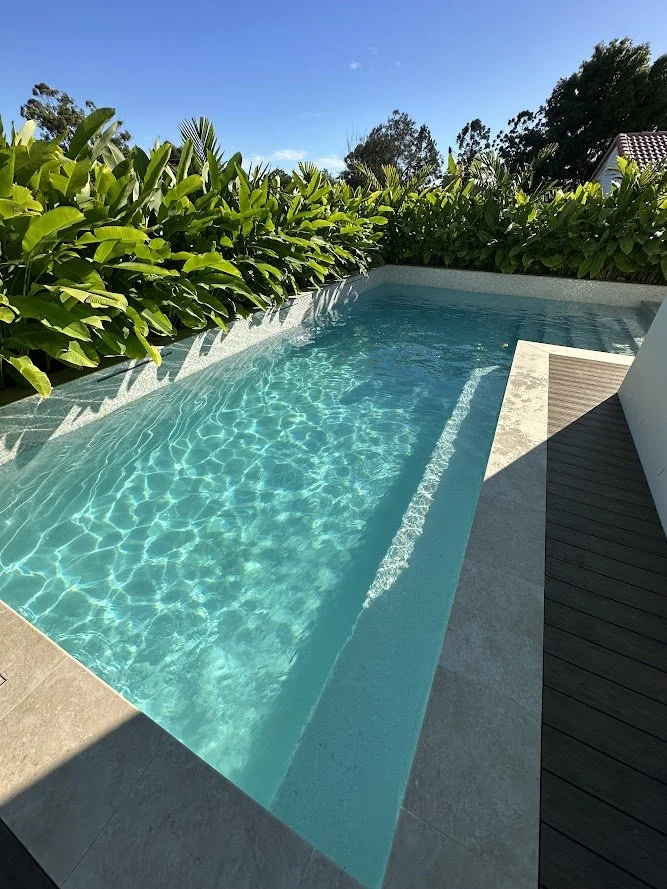 A small rectangular swimming pool with crystal clear water surrounded by green tropical plants, a wooden deck, and a bright blue sky overhead.