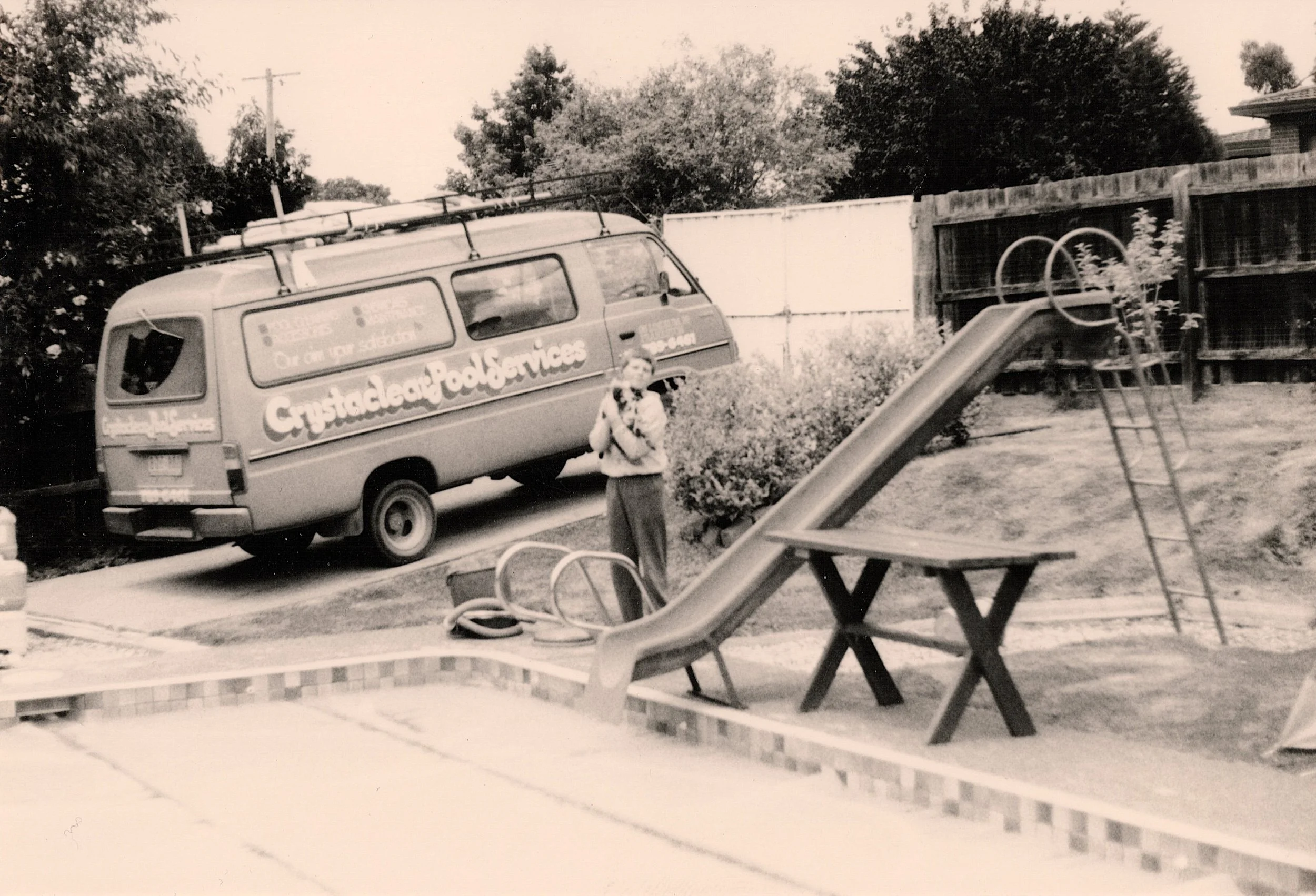 Black and white photo of a van parked in a yard with trees in the background. The van has signage advertising crystaclear pool services. There is a young Nick Briscoe standing next to an old slide and covered swimming pool visible in the yard.