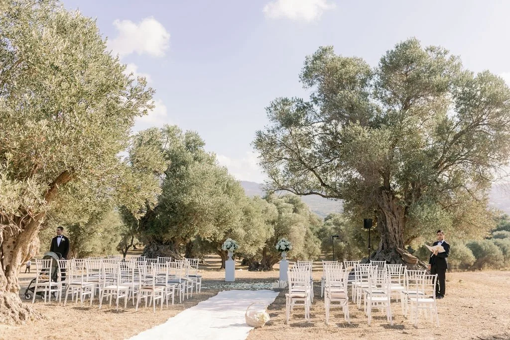 Outdoor wedding ceremony setup with white chairs on either side of a white aisle, large trees, and two people in formal attire.
