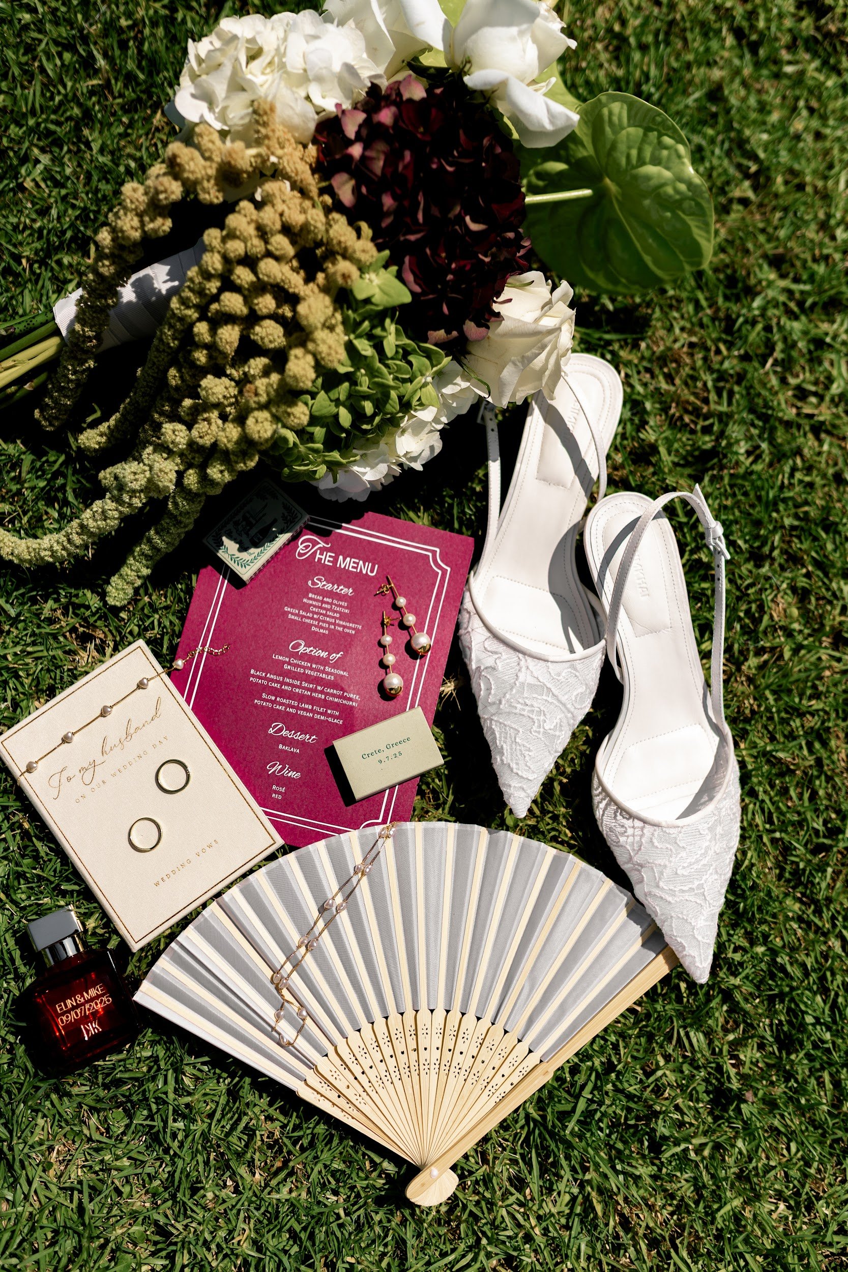 Flowers, accessories, and wedding items arranged on grass, including white high heels, a fan, jewelry, a menu, wedding rings, perfume, and decorative greenery.