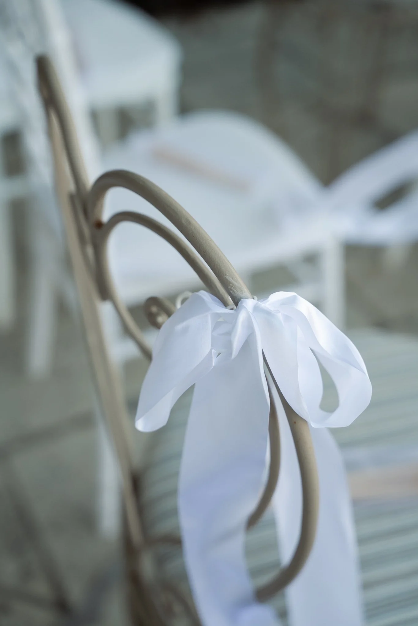 Close-up of white chair with a white ribbon tied to its metal frame, likely part of wedding or event decoration.