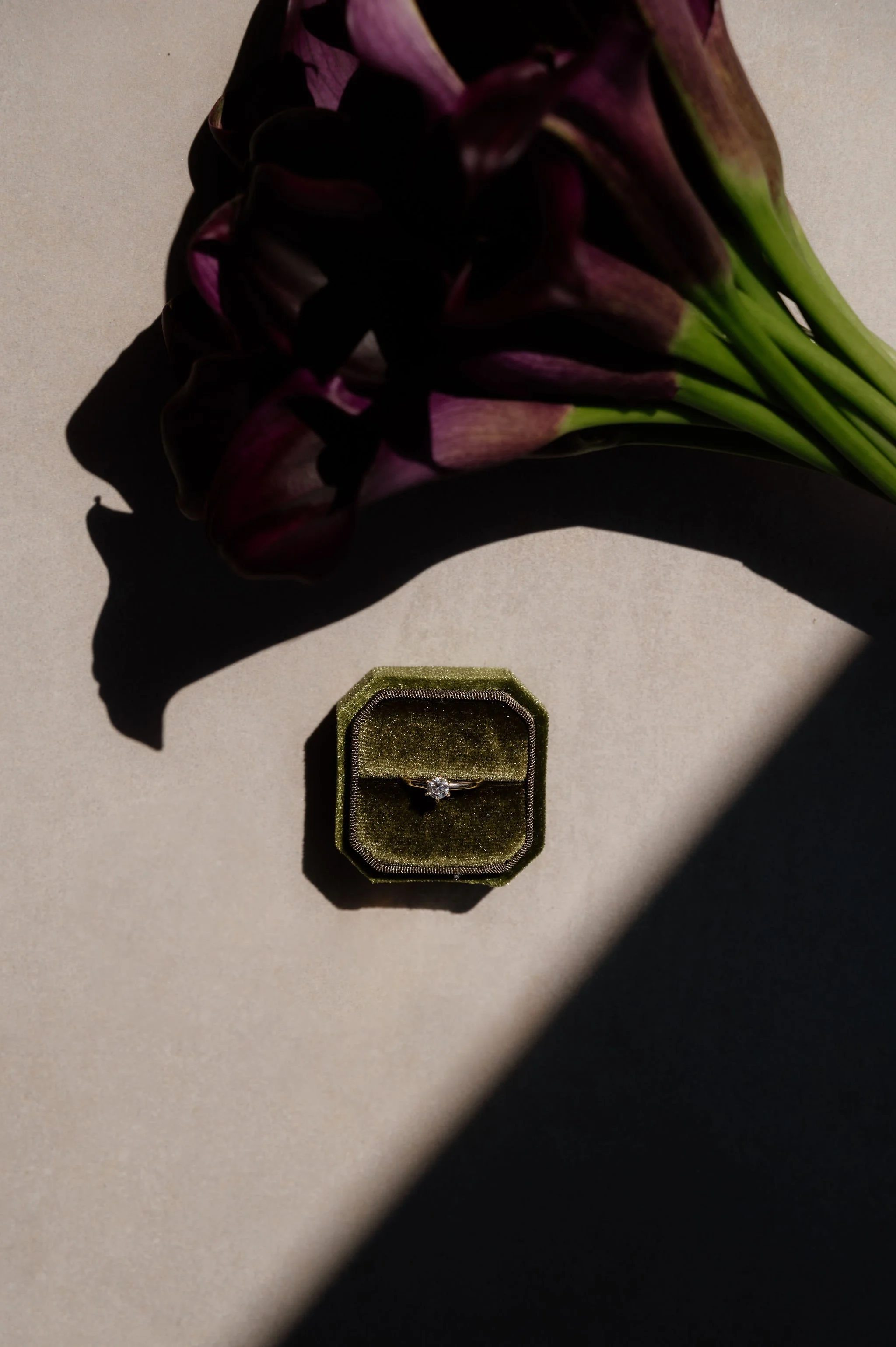 A small green velvet ring box contains a diamond engagement ring, placed on a light-colored surface with a deep purple flower next to it, casting shadows.
