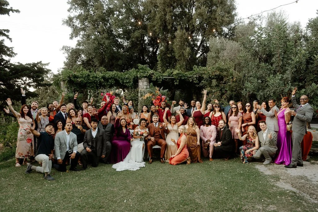 Large group of people at a wedding celebration outdoors, with many smiling and raising their hands, surrounded by trees and greenery.