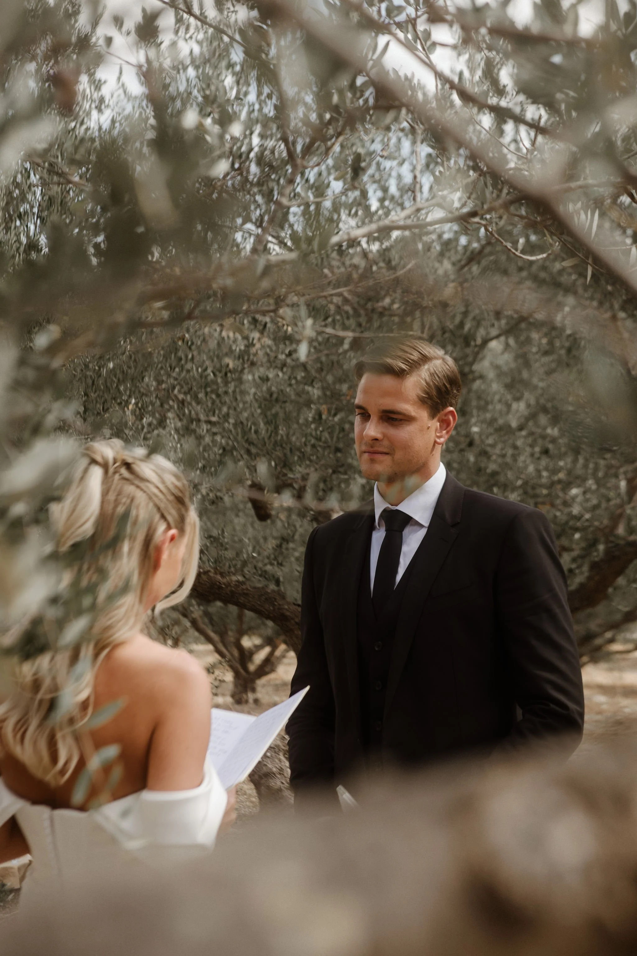 A man in a black suit and tie standing outdoors, with a woman reading from a paper in front of him, surrounded by trees and foliage.