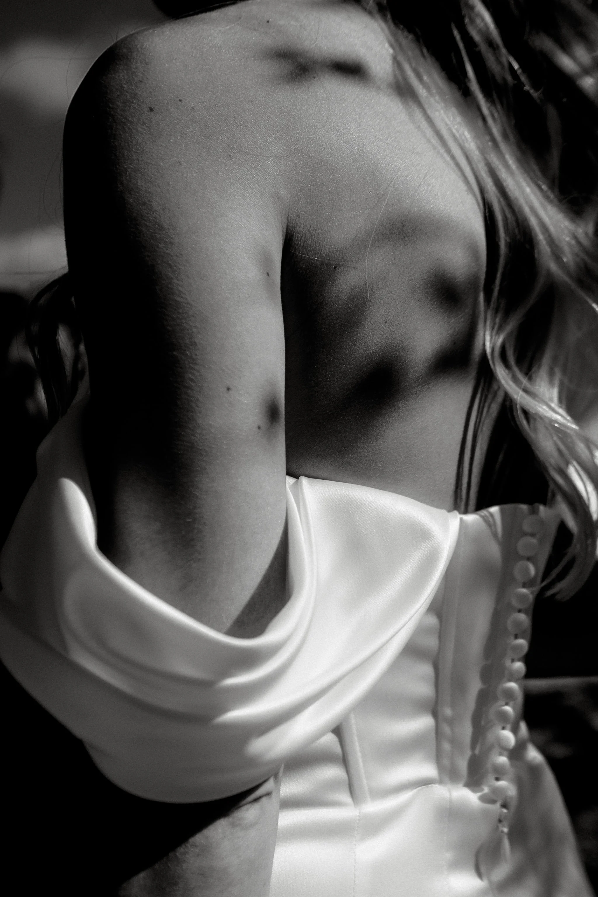 Close-up black-and-white photo of a woman’s shoulder, upper arm, and part of her face with long hair, wearing a satin garment with buttons.