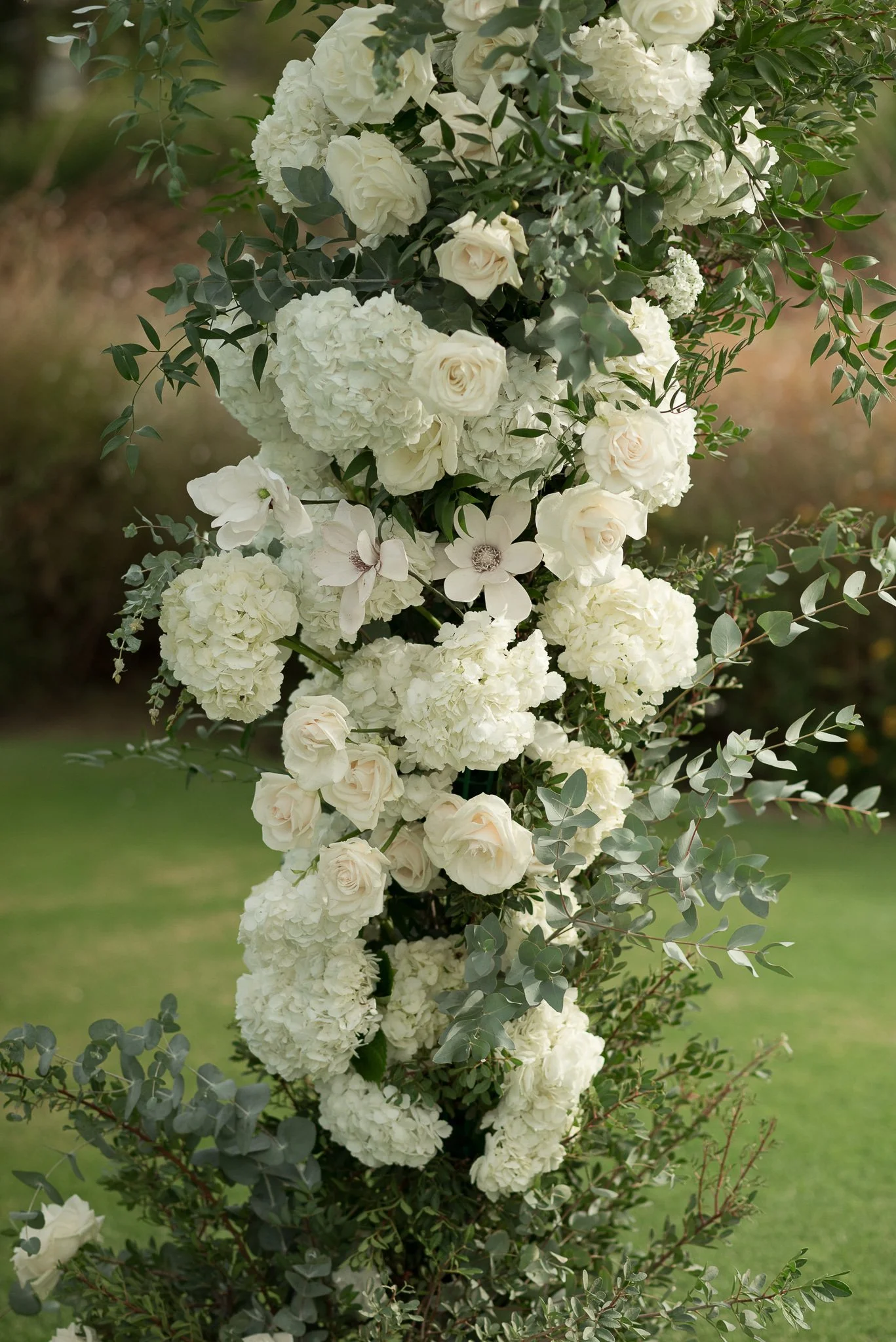 A vertical arrangement of white flowers, including roses, hydrangeas, and other blossoms, with green foliage in the background.