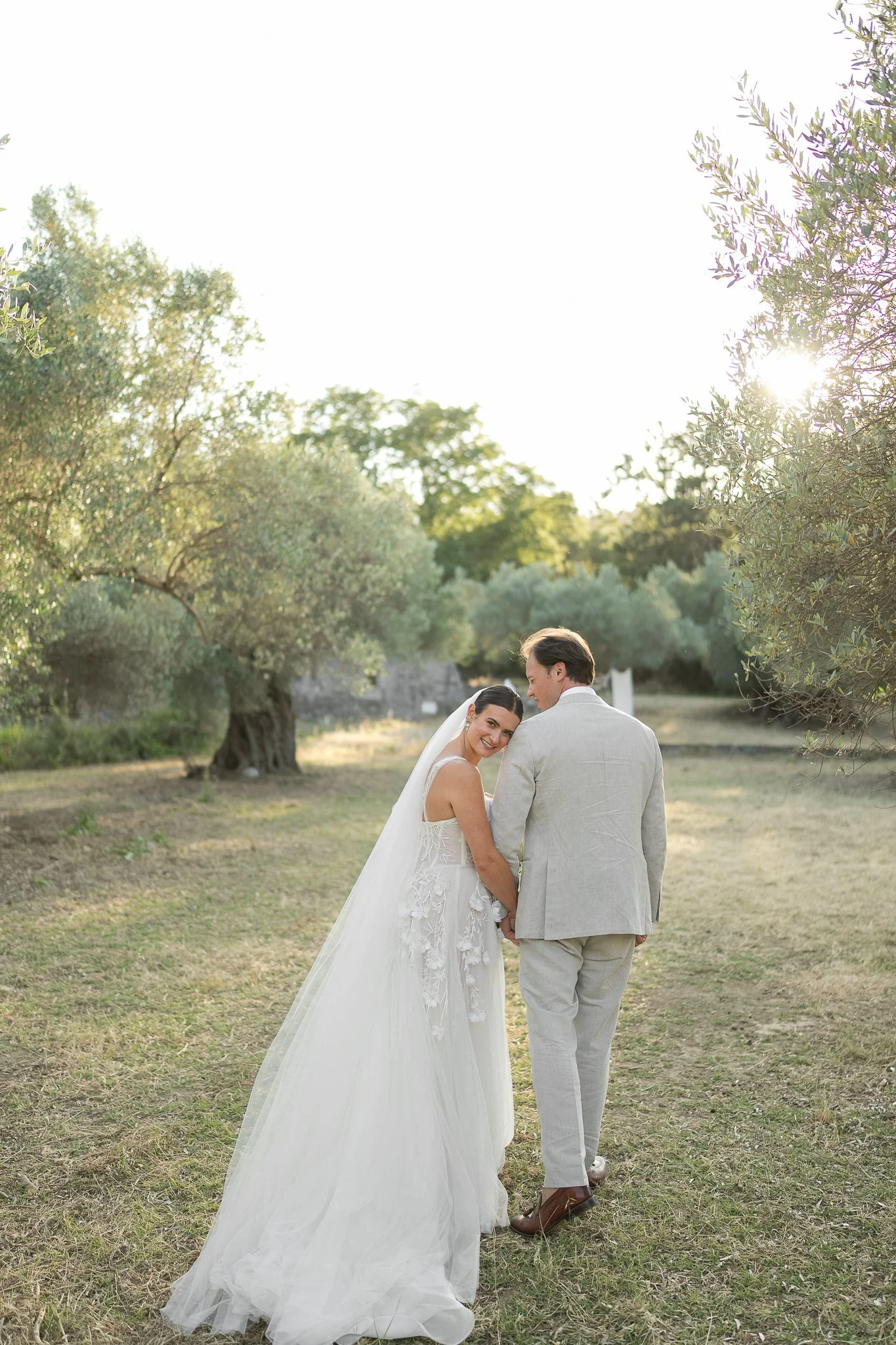 A bride and groom holding hands and smiling at each other outdoors during sunset, surrounded by trees.