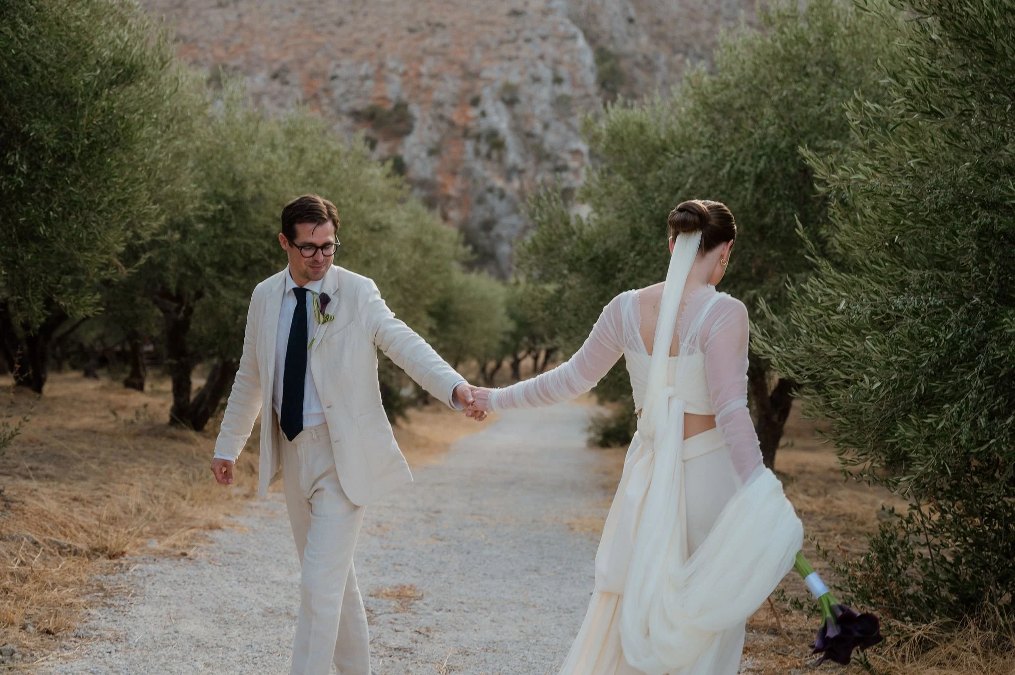 A newlywed couple walks hand in hand along a dirt path lined with green trees in a rural outdoor setting, with a rocky hillside in the background. The groom is wearing a cream-colored suit with a dark tie and glasses, and the bride is dressed in a light-colored, semi-sheer wedding gown with a long veil and holding a dark purple flower bouquet.