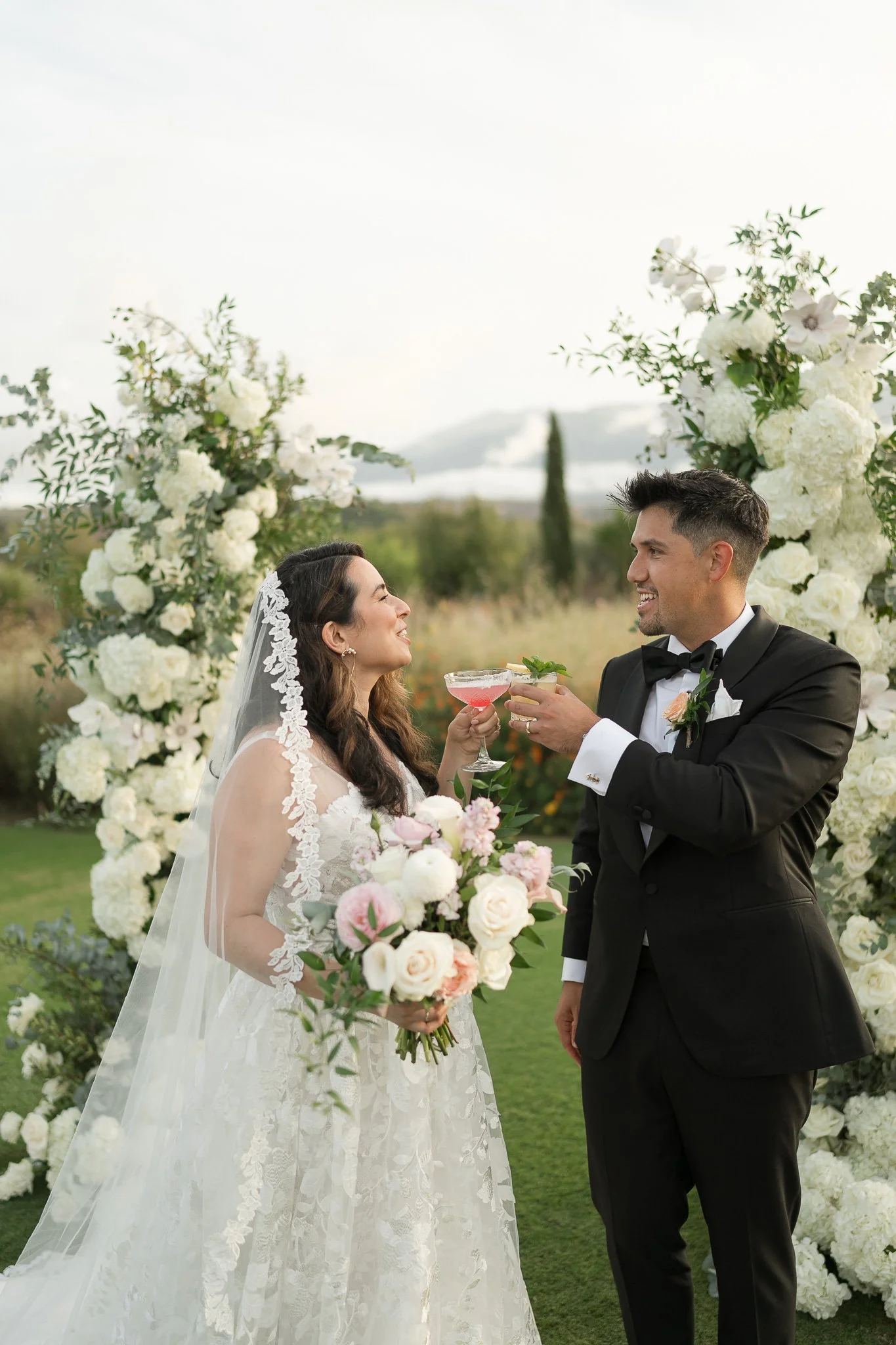 A bride and groom standing outdoors during their wedding ceremony, holding glasses and smiling at each other. The bride is wearing a white lace wedding dress and veil, holding a bouquet of pink and white roses. The groom is in a black tuxedo with a b