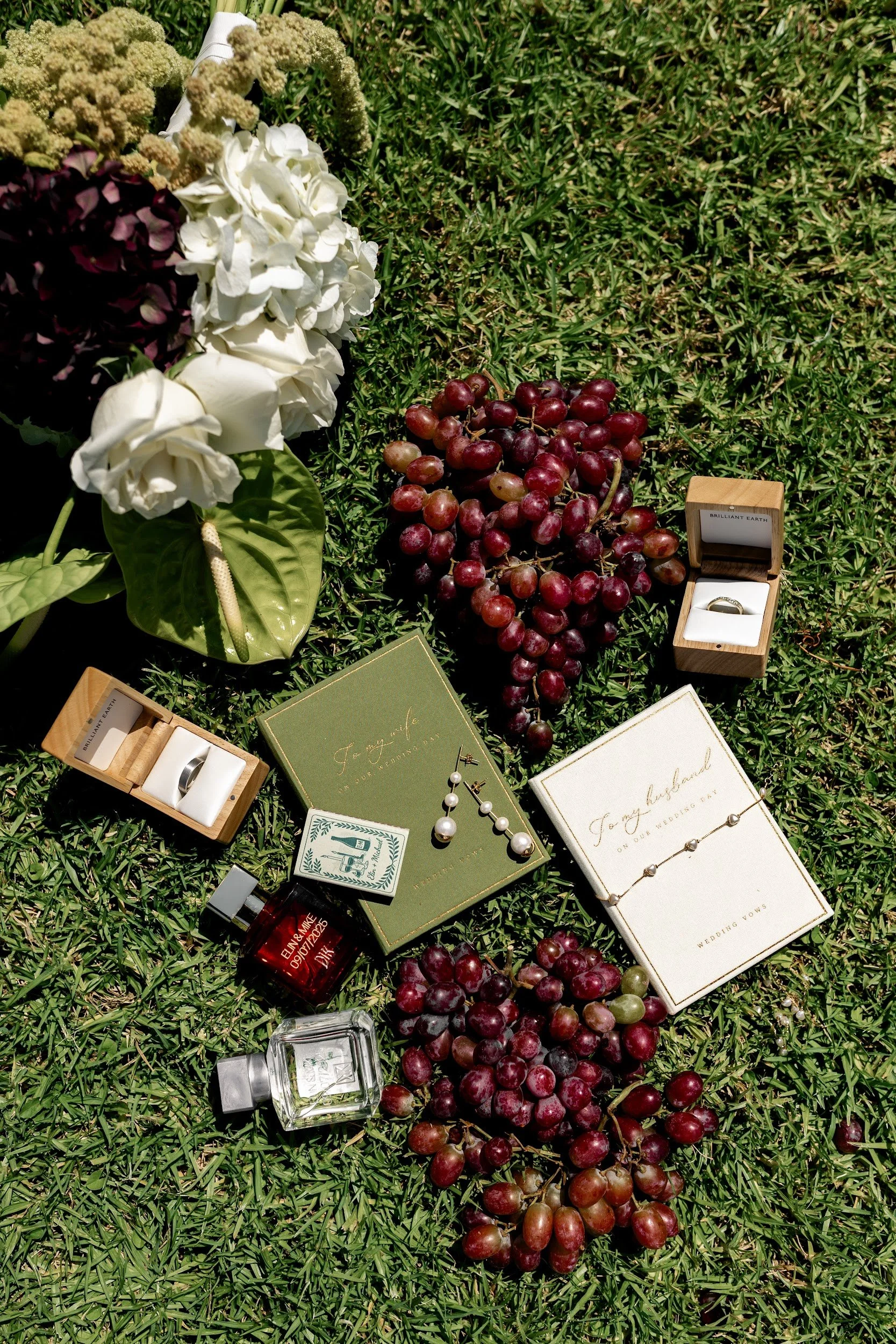A wedding flat lay on green grass featuring flower arrangements, bunches of red and green grapes, two wedding rings in boxes, two sets of wedding vows, a perfume bottle, and a small jar bottle.