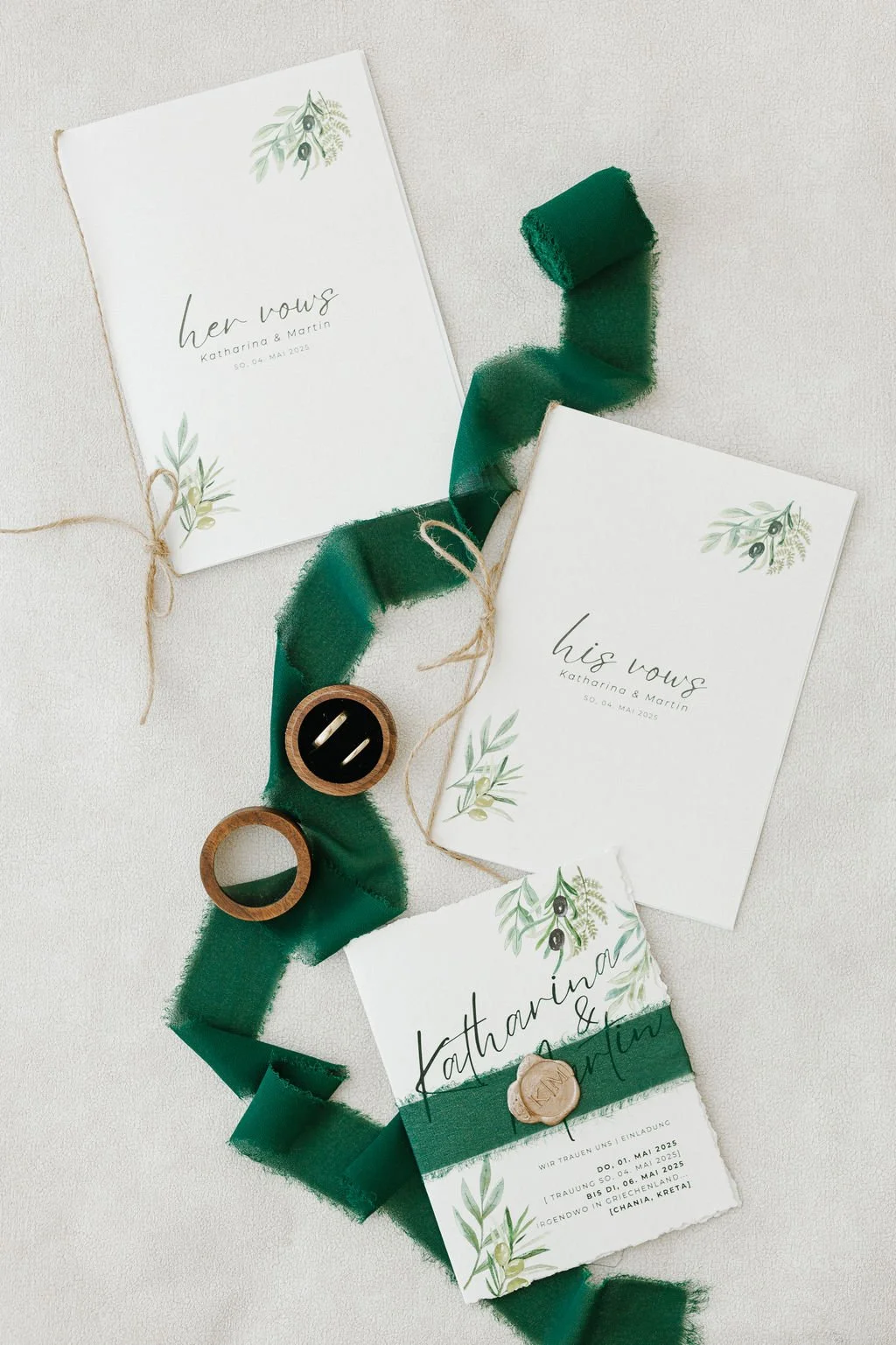 Invitation cards for Katharina and Martin's wedding, decorated with green leaves and black olives, with a green ribbon, a small wooden wax seal, and two wooden rings placed on a light surface.
